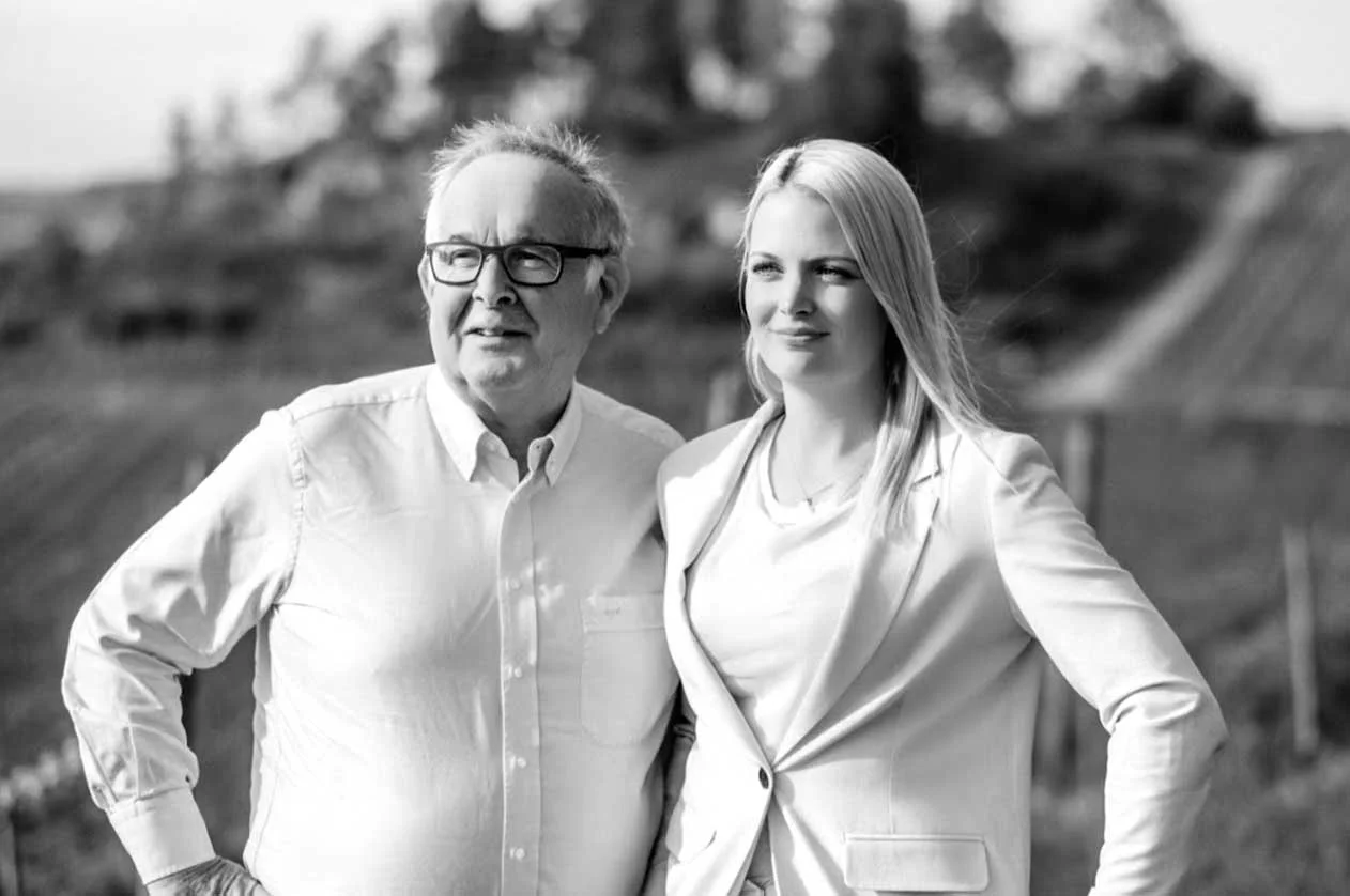 A black and white photo of an older man with glasses and a woman, both dressed in light-colored clothing, standing outdoors with a blurred natural landscape in the background.