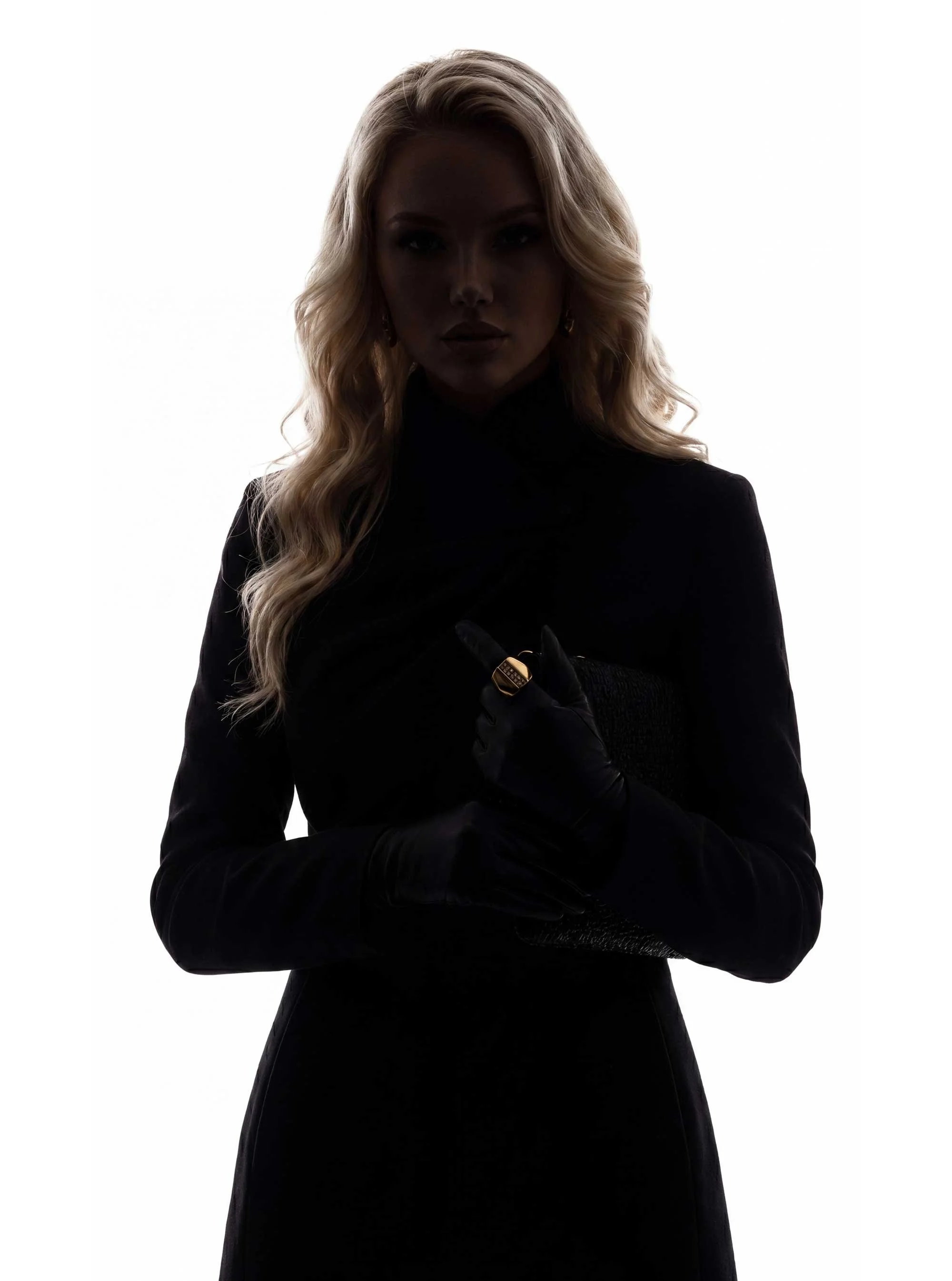 A woman with long, wavy blonde hair wearing a dark outfit and gloves, standing against a plain white background.