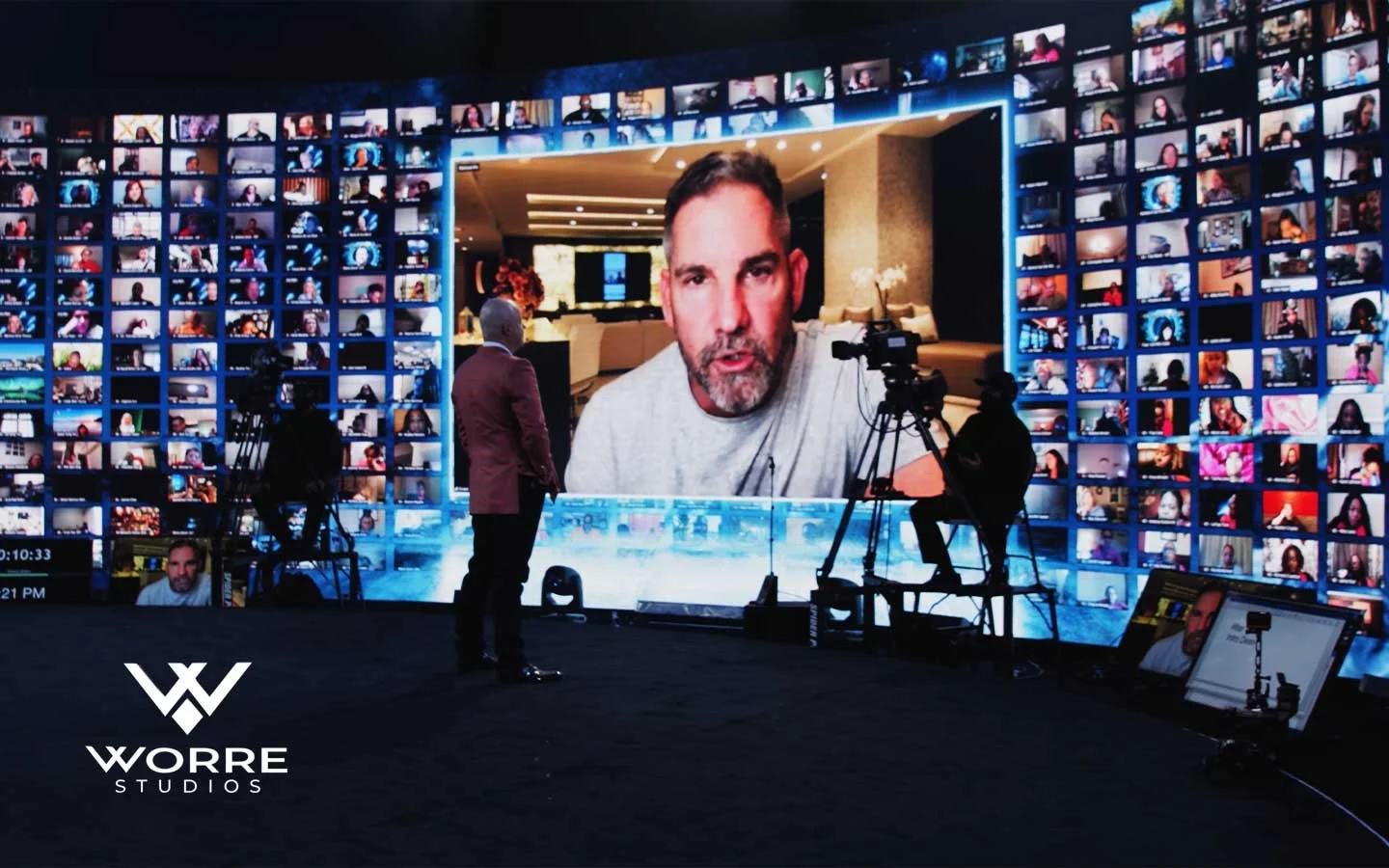 A large screen displaying a man with dark hair and a beard speaking to the camera, surrounded by numerous smaller screens of virtual participants, with crew members setting up equipment in front of the stage at WORRE Studios.