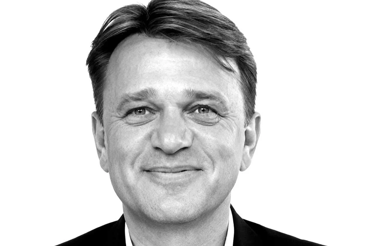 Black and white headshot of a middle-aged man with short, combed-back hair, smiling, wearing a dark suit and white shirt.