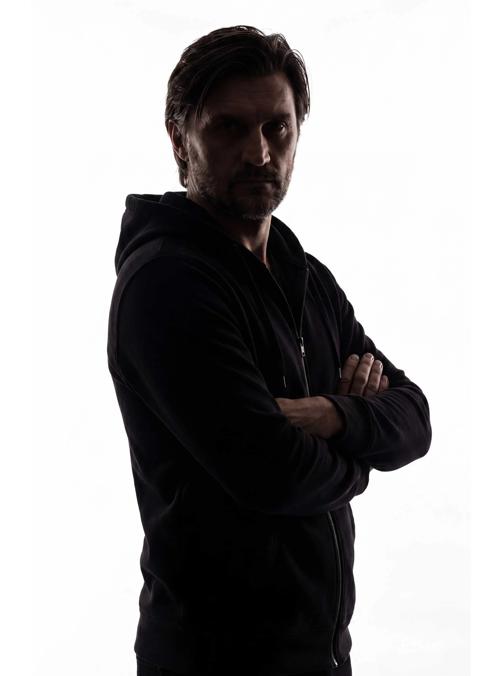 A man with dark hair and a beard, wearing a black hoodie, stands with arms crossed against a white background, looking directly at the camera.