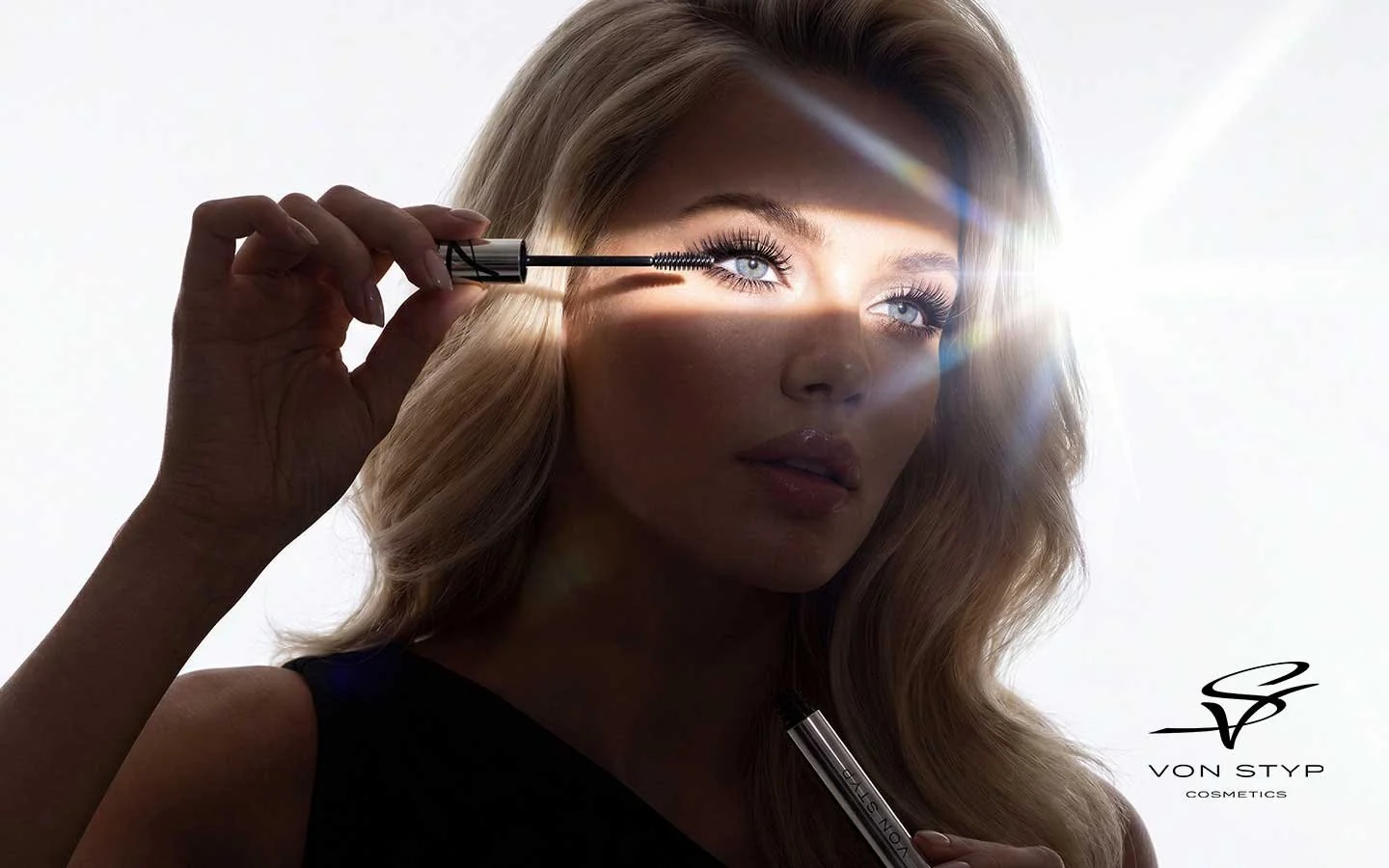 A woman with blonde hair and blue eyes applying mascara with a brush while holding a lipstick in her hand. There's a bright light source behind her, causing a lens flare. The image features the Von StyP Cosmetics logo in the bottom right corner.
