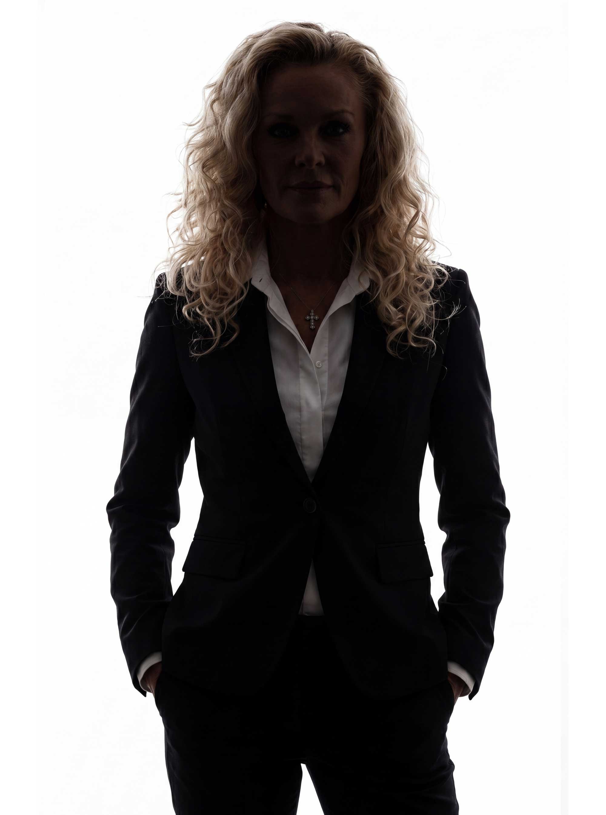 A woman with curly blonde hair standing against a bright white background, dressed in a black suit with a white shirt and a cross necklace.