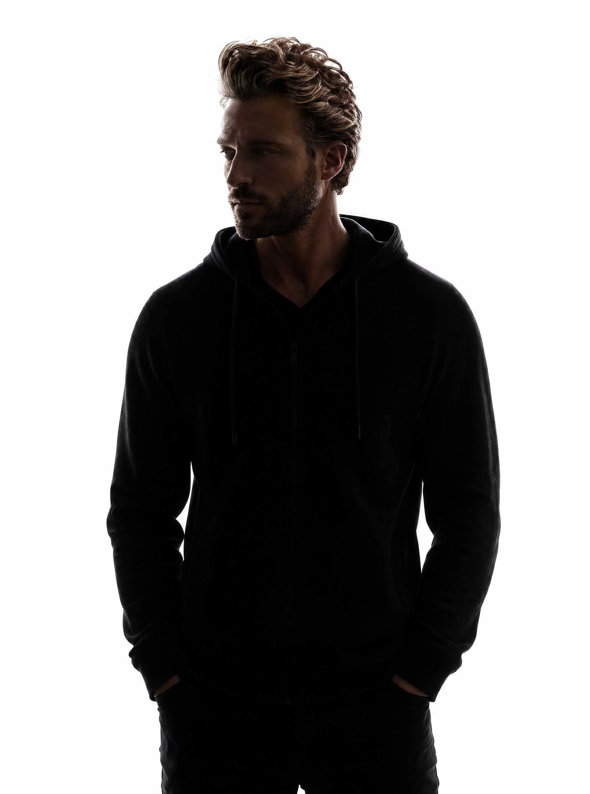 A man with brown hair and stubble wearing a black hoodie stands against a white background, looking to the side.