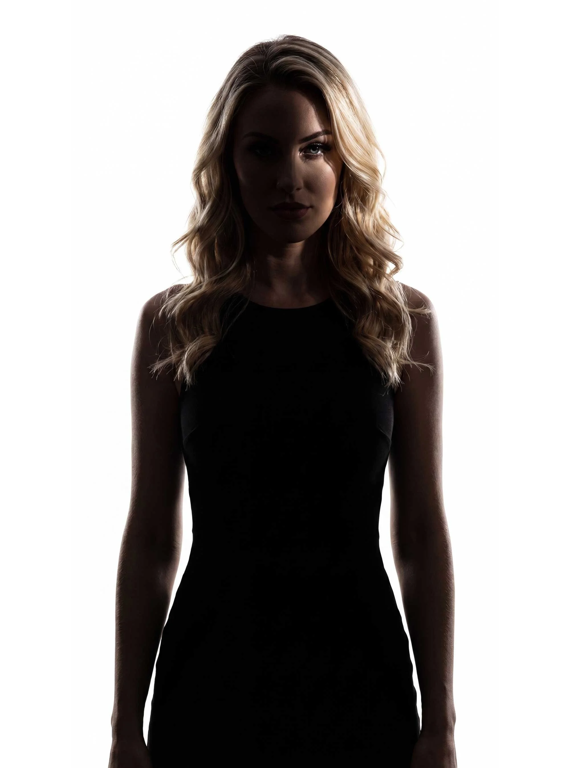A woman with long, wavy blonde hair wearing a sleeveless black top, standing against a white background, with lighting creating a high contrast silhouette effect.