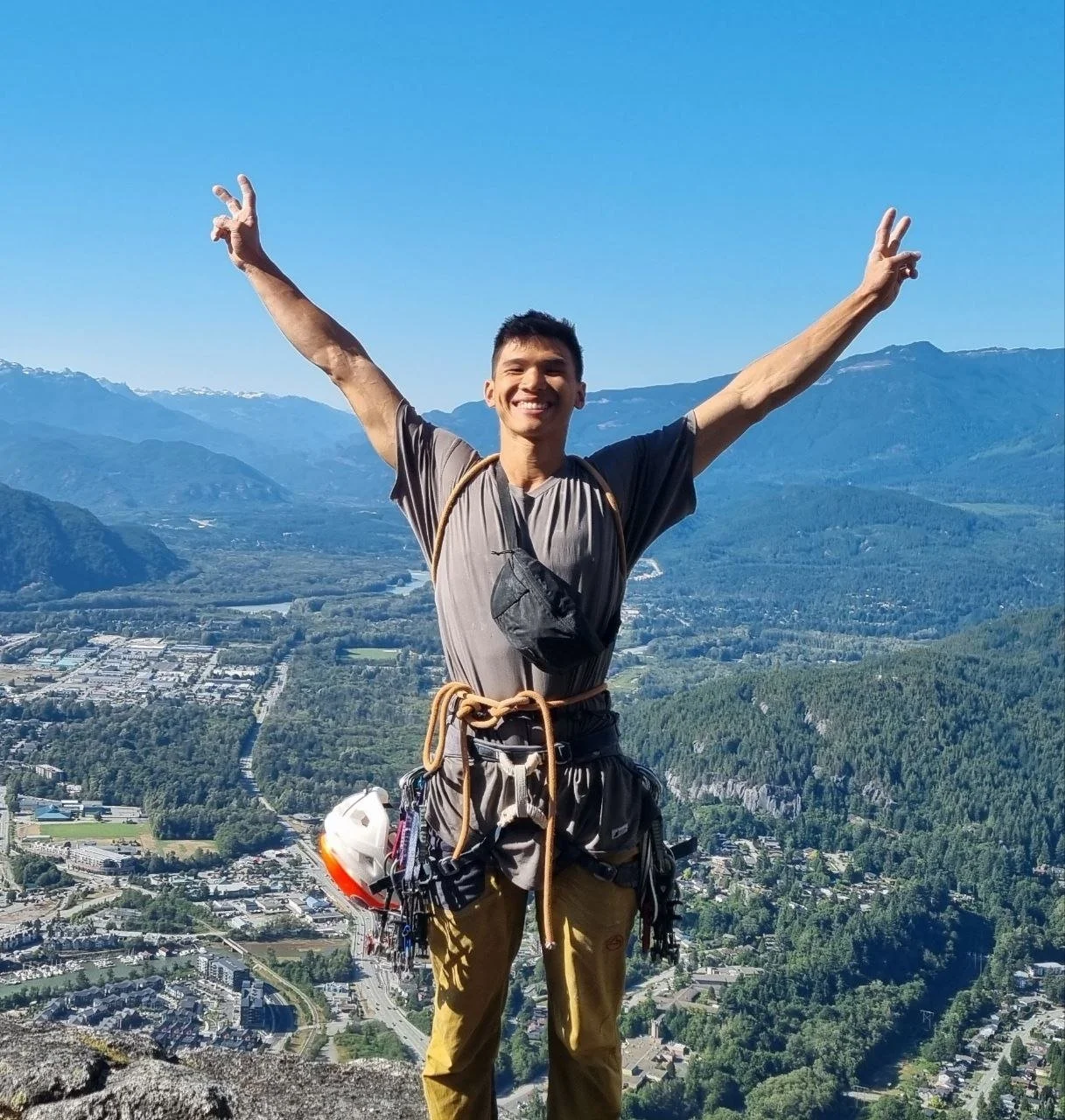 A smiling man with arms outstretched standing on a hilltop with a view of a valley, mountains in the background, and a clear blue sky.