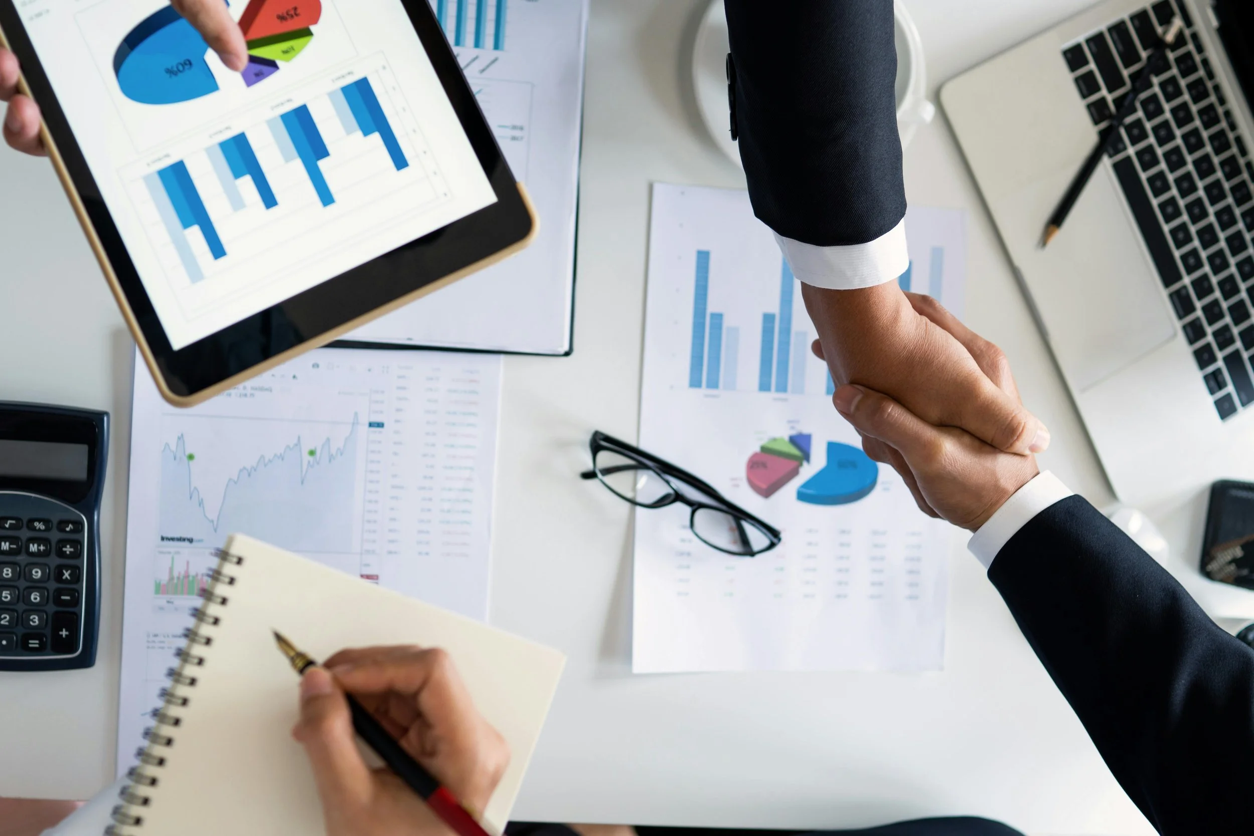 Personi che tengono per mano durante una riunione di lavoro con grafici, report e laptop su una scrivania.