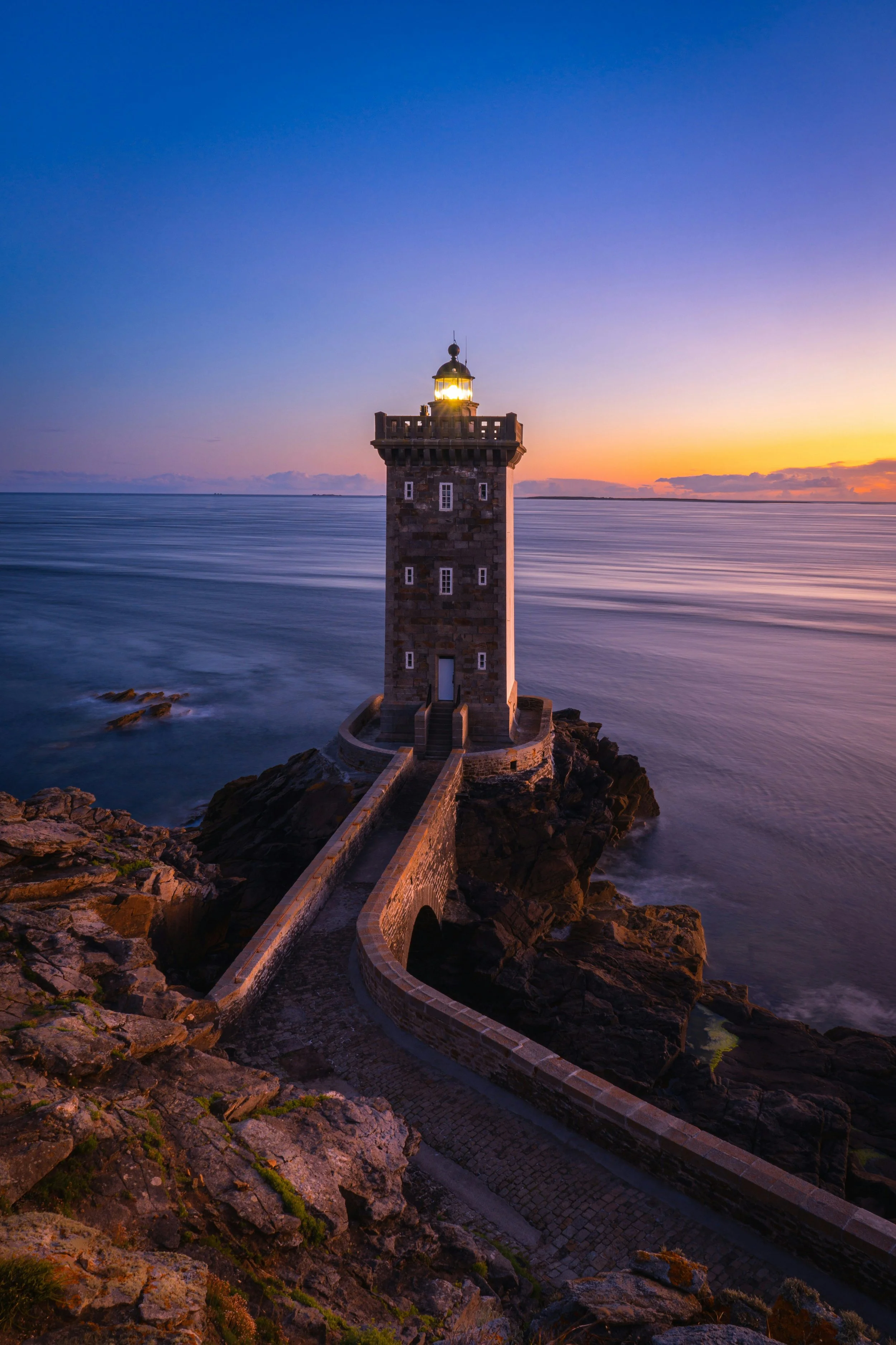 Faro a Torre di San Juan de Gaztelugatxe vista durante il tramonto, con il cielo blu e sfumato di arancione, sul mare calmo.