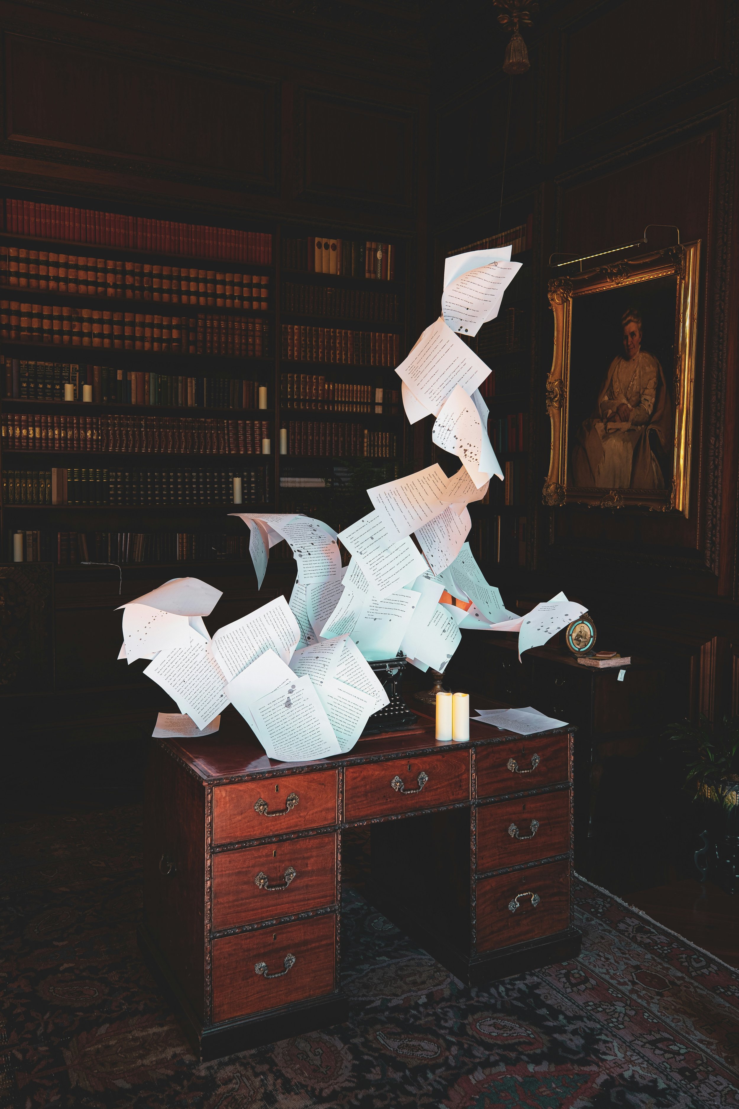 Una stanza con pareti di libri e un dipinto di una donna seduta in un quadro dorato. Al centro, una scrivania con cassetti di legno, coperta da molte pagine di carta che sembrano volare nell'aria, creando un effetto di movimento.