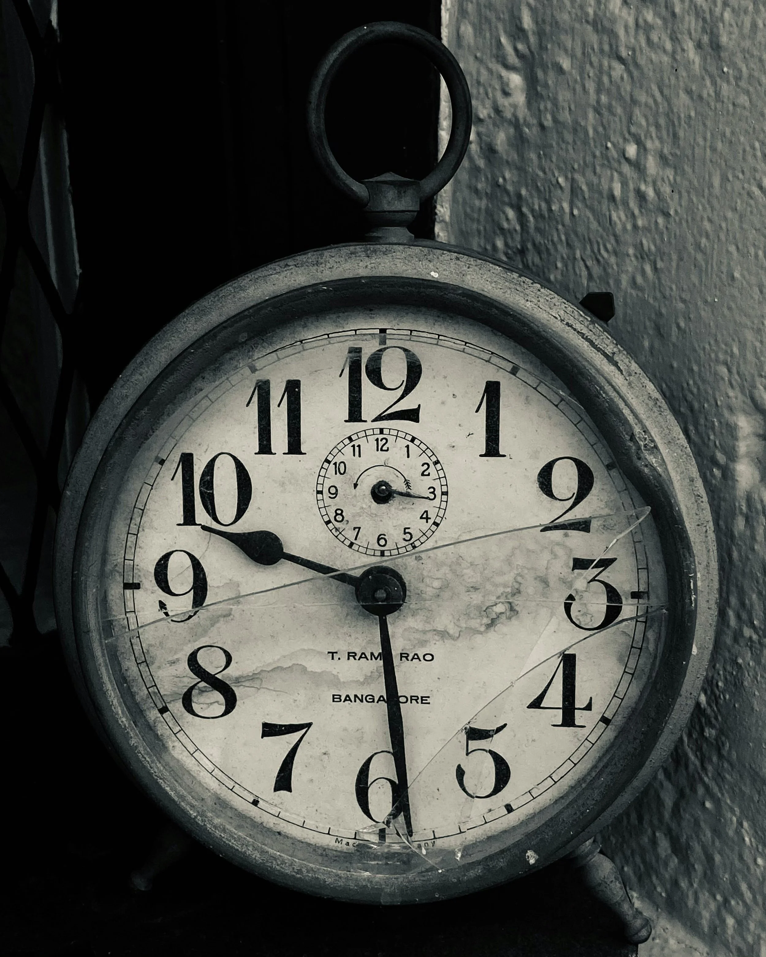 Orologio antico rotto con grande quadrante bianco, numeri neri e due lancette nere, appoggiato su una superficie nera con muro di pietra sullo sfondo.
