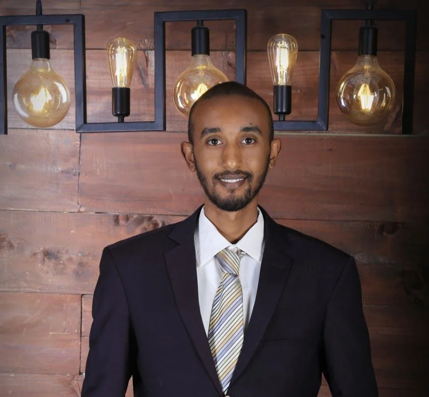 A man in a dark suit, white shirt, and striped tie standing in front of a wall with wooden panels and four hanging Edison-style light bulbs.