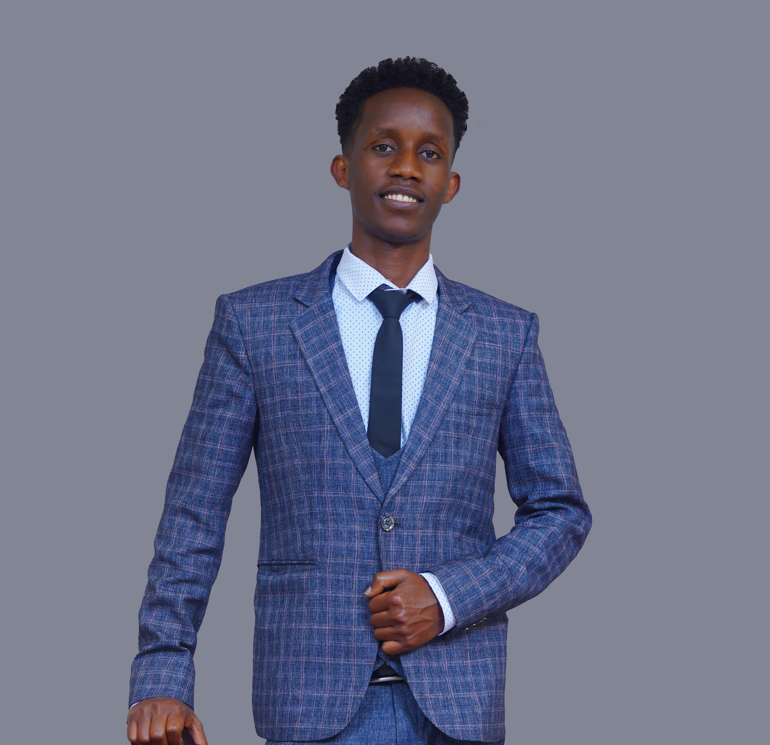 A young man wearing a checked blazer, white shirt, and dark tie, standing against a gray background, smiling confidently.