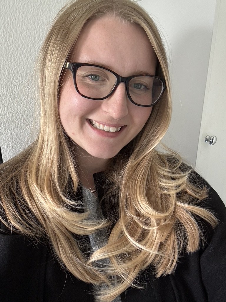 A woman with long, wavy blonde hair, wearing glasses and a black jacket, smiling at the camera indoors.