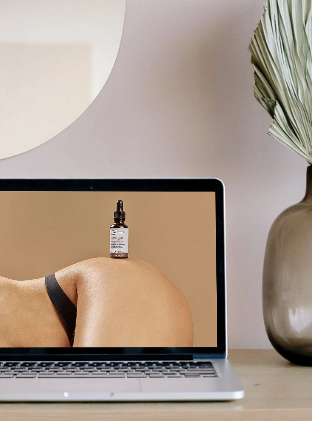 A laptop on a desk shows a close-up of a woman's leg with black underwear, a small bottle of facial serum placed on her knee, and a desktop background with the serum bottle. A vase with large, dried palm leaves is on the right side, and a wall-mounted lamp is visible in the background.