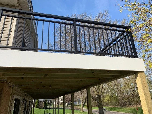 View of a newly constructed deck with black metal railing, supported by wooden posts, attached to a house with a backyard featuring trees and a lawn.