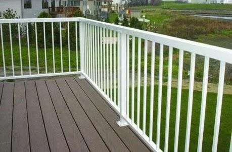 View of a white metal balcony railing on a wooden deck, overlooking a grassy area and some houses in the distance.