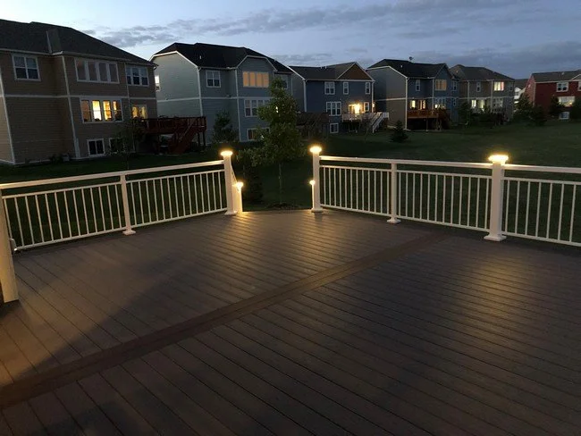 A wooden deck with white railing and built-in lights overlooking a suburban neighborhood at dusk, with multiple houses and a green lawn.