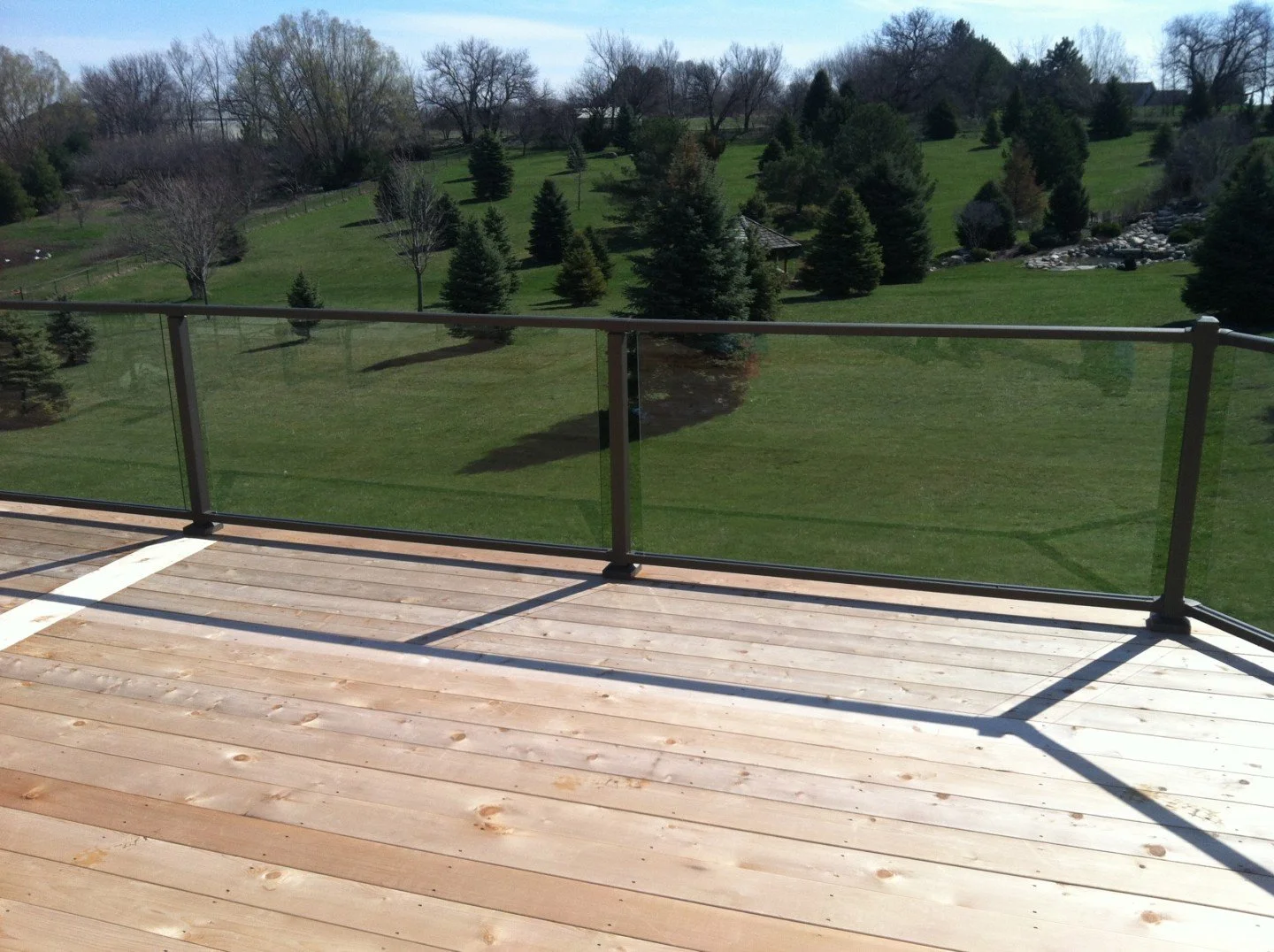 Wooden deck with glass railing overlooking a grassy, tree-filled landscape with some rocks and distant houses under a partly cloudy sky.