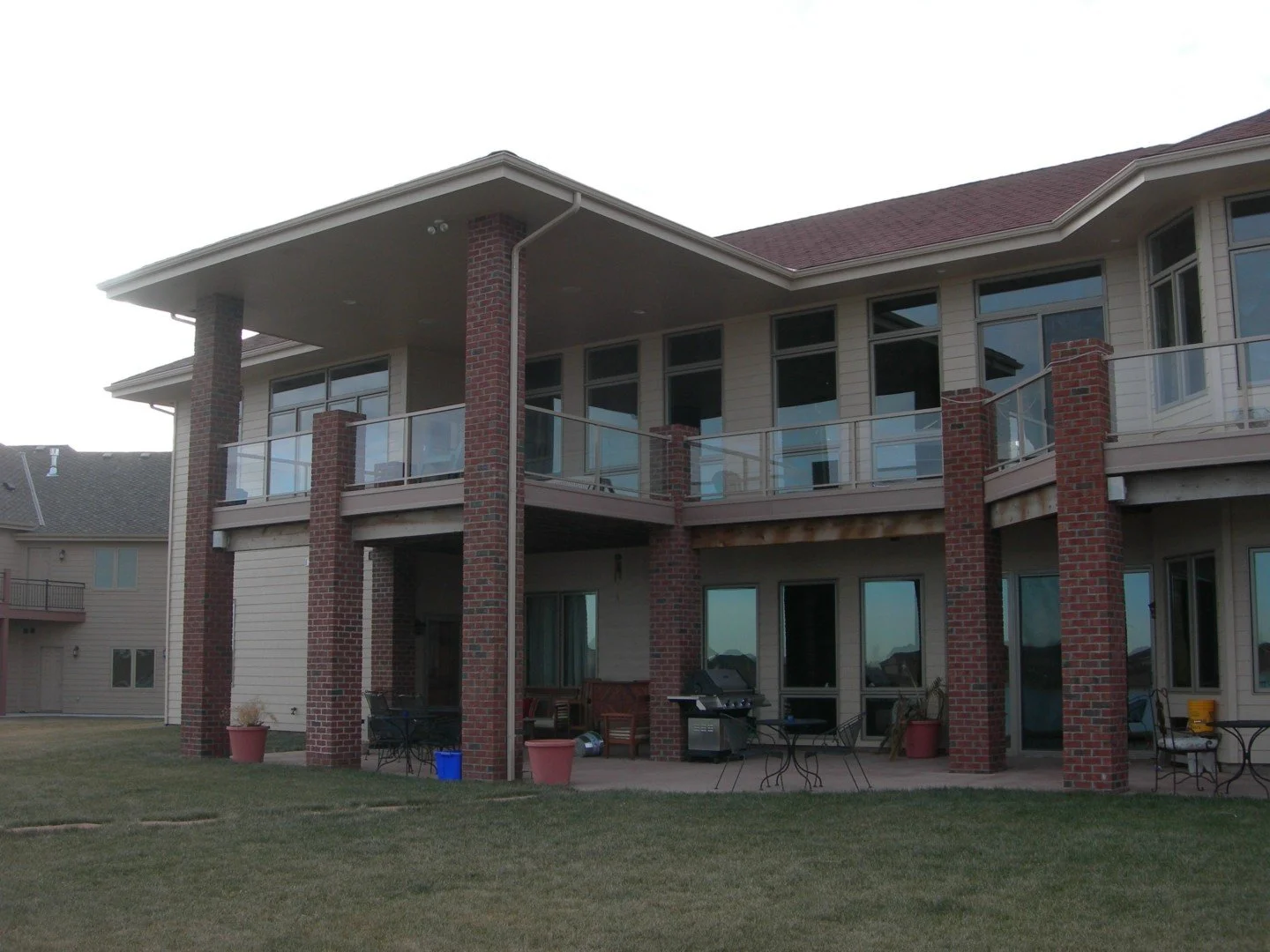 A two-story house with a brick and beige siding exterior, large windows, and a balcony with glass railing. The backyard features a patio with outdoor furniture, potted plants, a barbecue grill, and a grassy lawn.