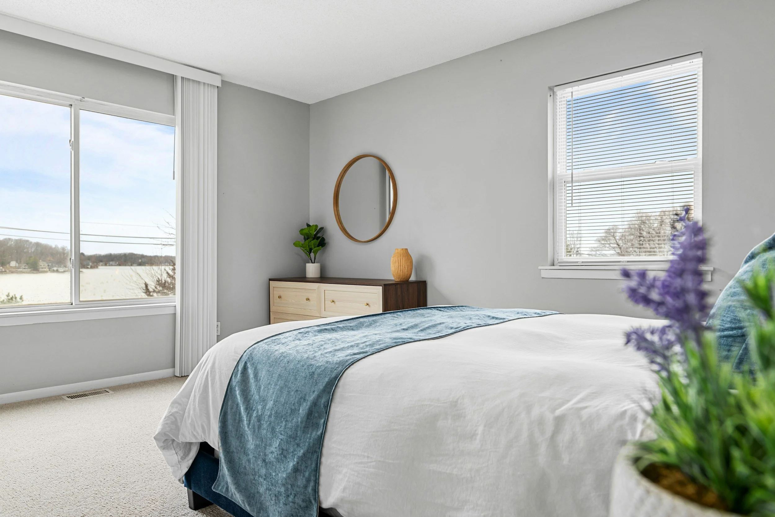 Bedroom with large window showing a blue sky and trees, light gray walls, white carpet, bed with white bedding and blue throw, wooden dresser with decorative items, and a small plant.
