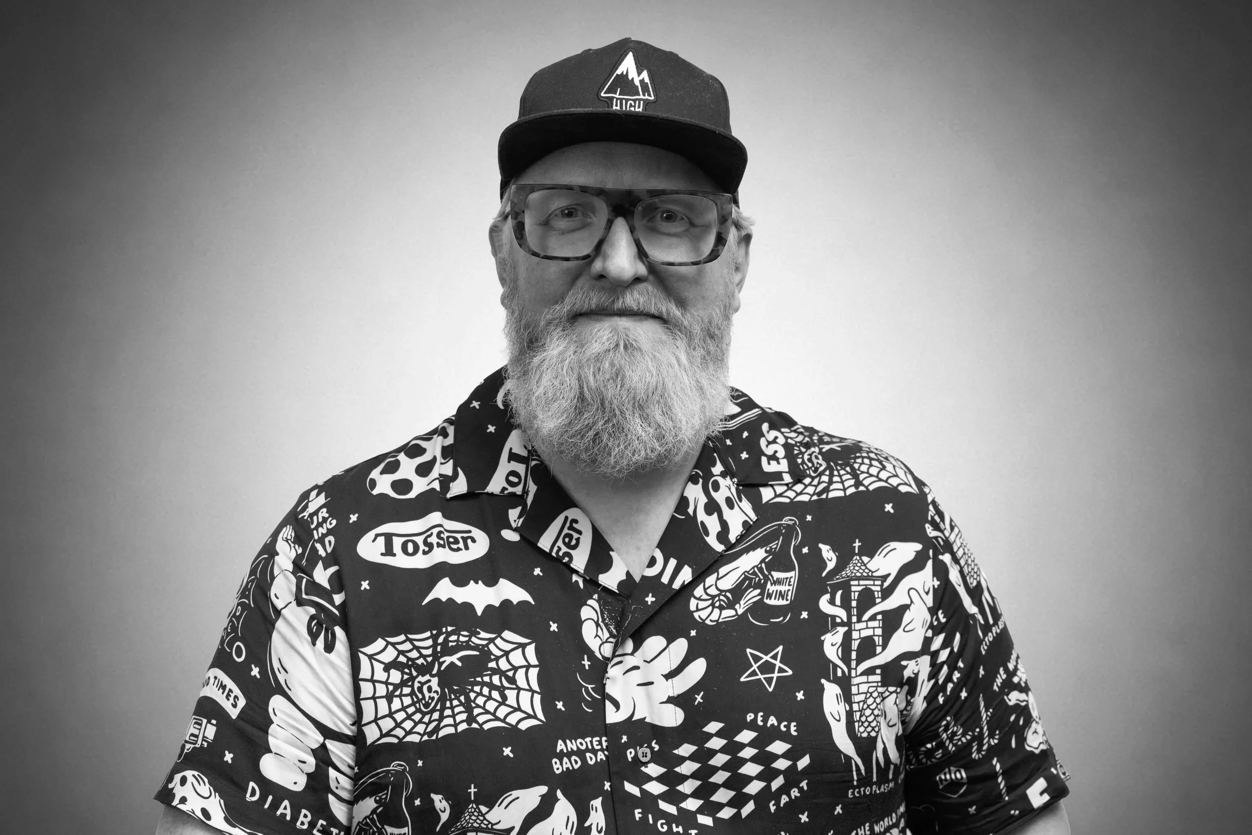 Kristjan Byfield: A man with glasses and a beard wearing a black baseball cap and a patterned shirt standing against a plain background.