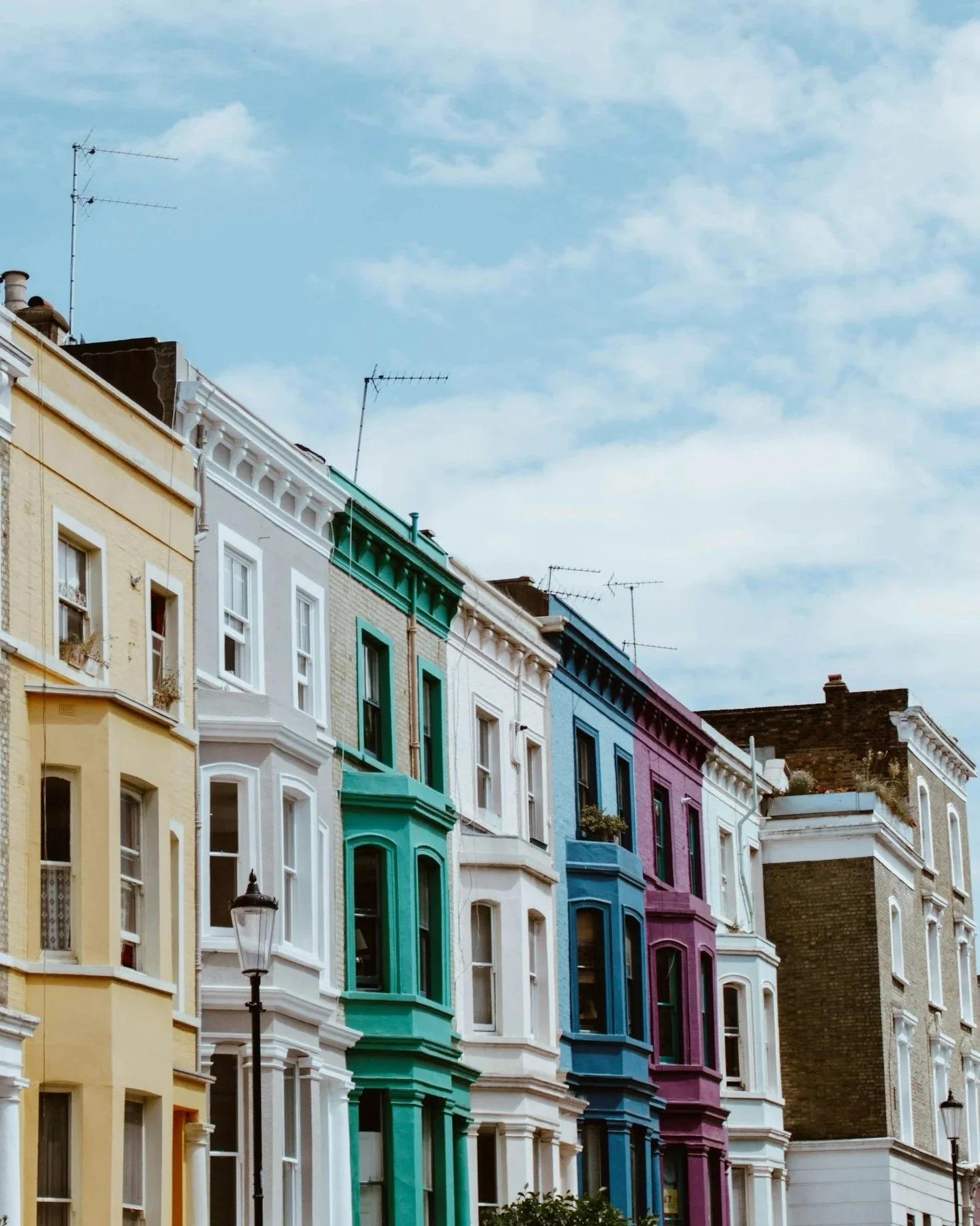 Colorful row of Victorian-style townhouses painted in pastel colors including yellow, green, white, blue, purple, and darker shades, with decorative trim and large windows, under a partly cloudy sky.