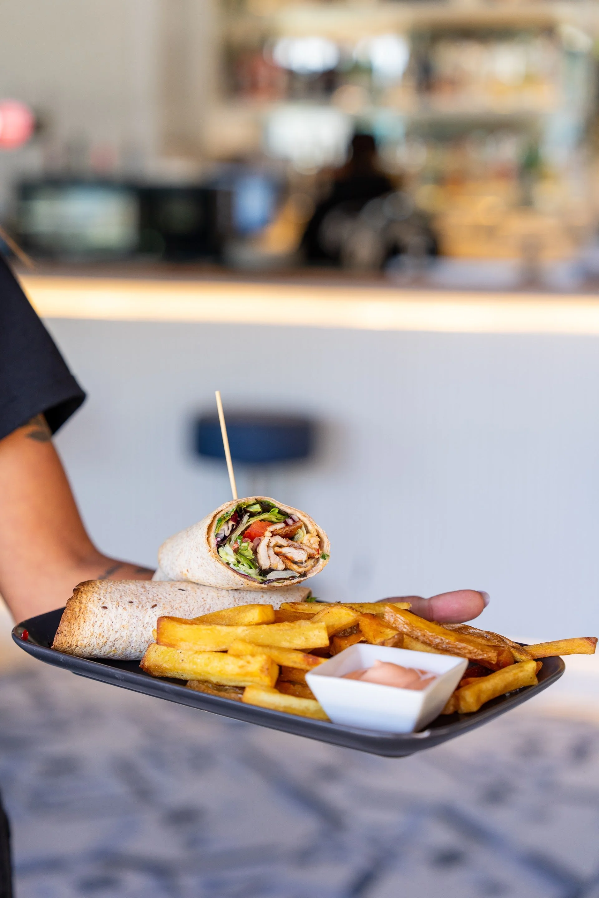 A person holding a black plate with French fries, a wrap filled with vegetables and grilled chicken, and a small dish of dipping sauce, in a restaurant setting.