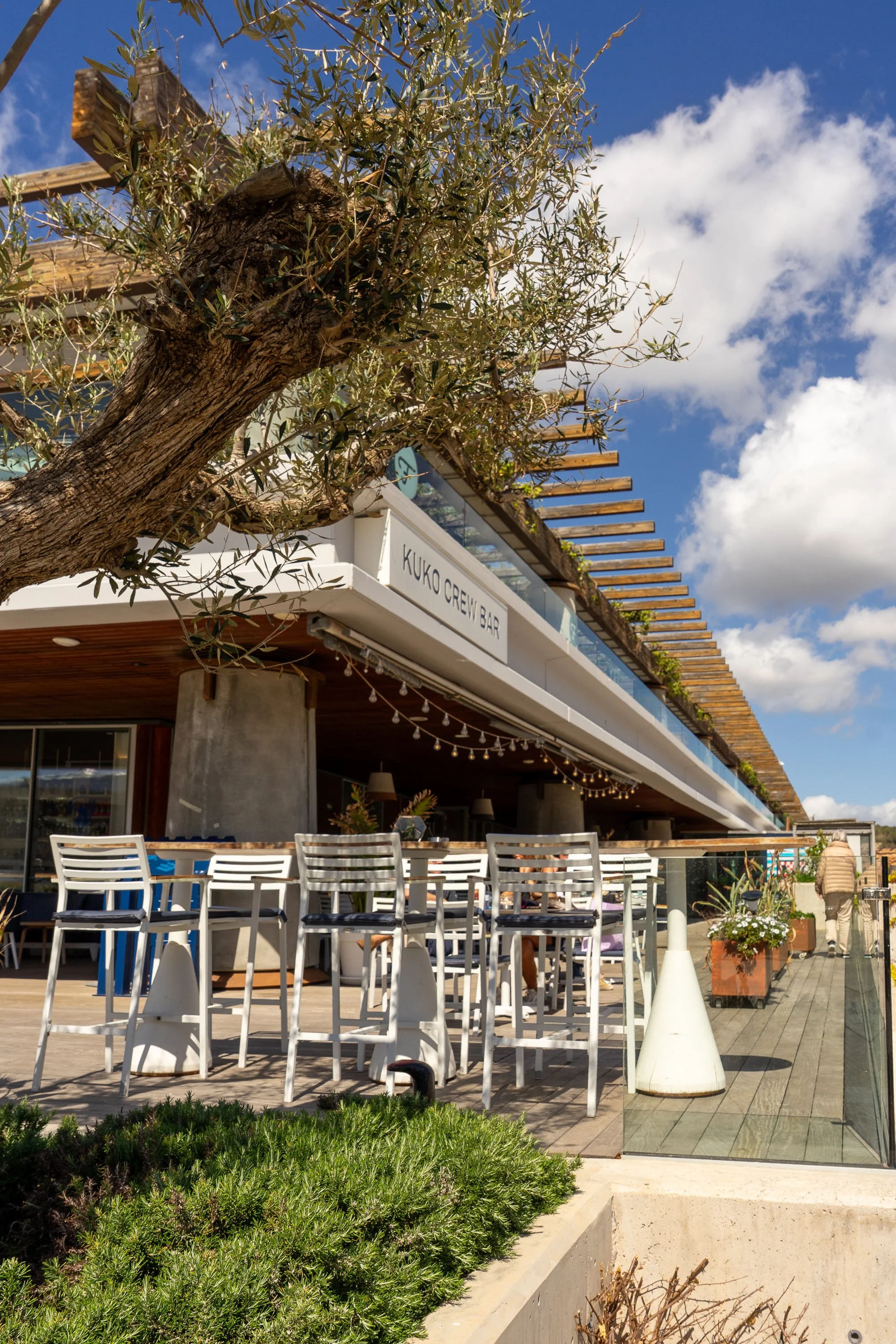 Outdoor seating area with white chairs, tables, umbrellas, and greenery in front of a modern building with a sign that reads 'KUKO CREW BAR,' under a partly cloudy sky.