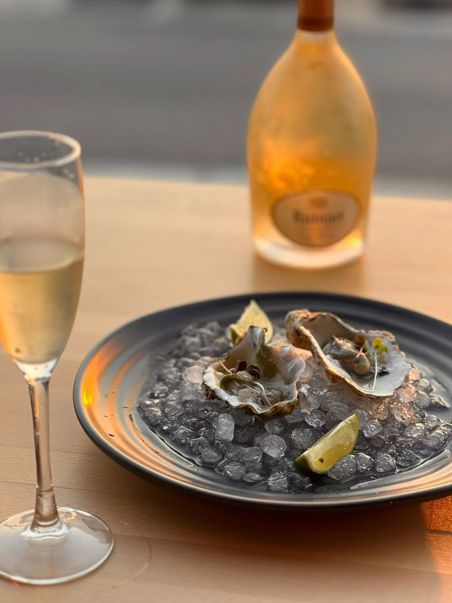 A glass of champagne and a plate of oysters on ice with lemon wedges, with a bottle of rosé wine in the background.