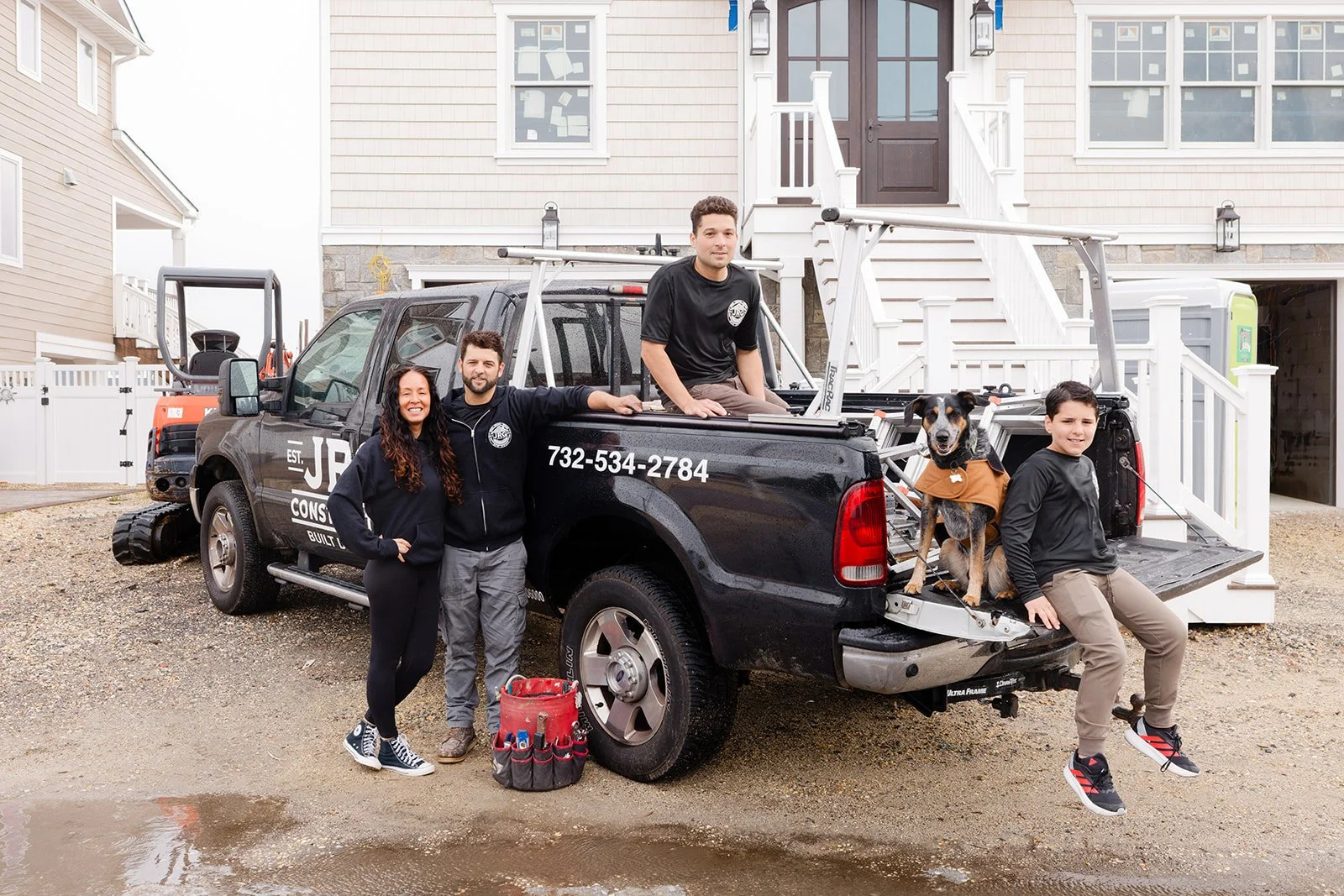 Five people and a dog pose in front of a utility truck outside a house. The truck has tools and a ladder, and the house has a white staircase. The group is smiling and dressed casually.