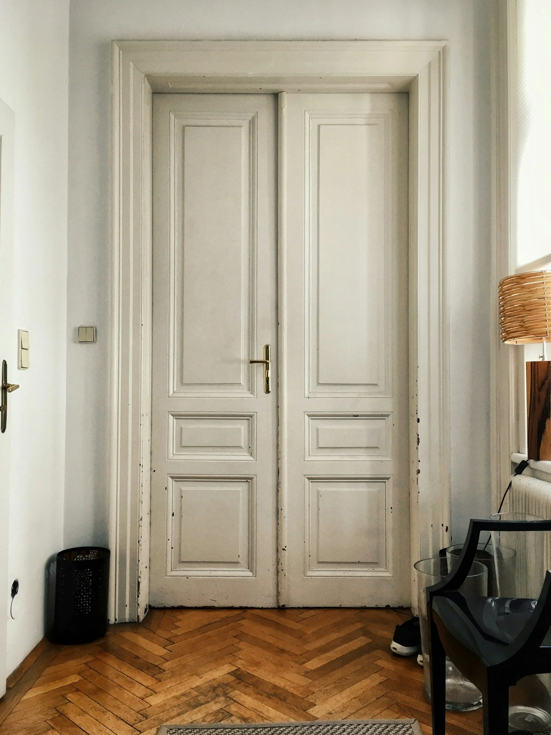 White double doors with molding details in a room with wooden herringbone flooring, a black plant pot on the left, a glass side table, and a black chair with shoes and a clear plastic container nearby.