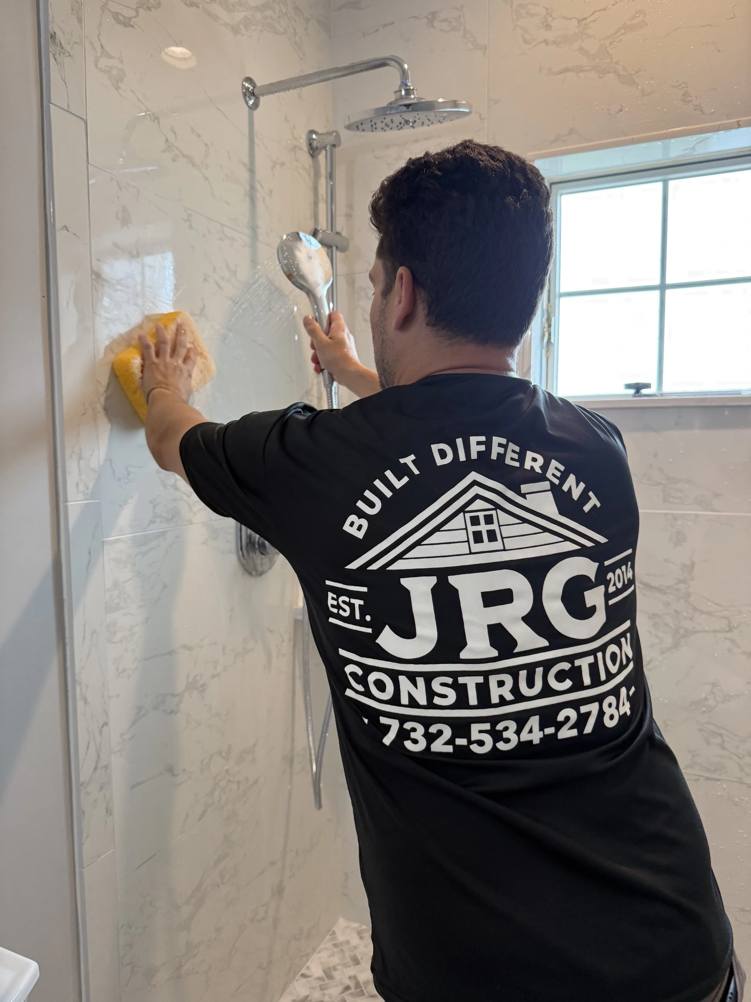 A man cleaning a shower wall with a yellow sponge, holding a showerhead in the other hand, in a modern bathroom with marble walls and a window.