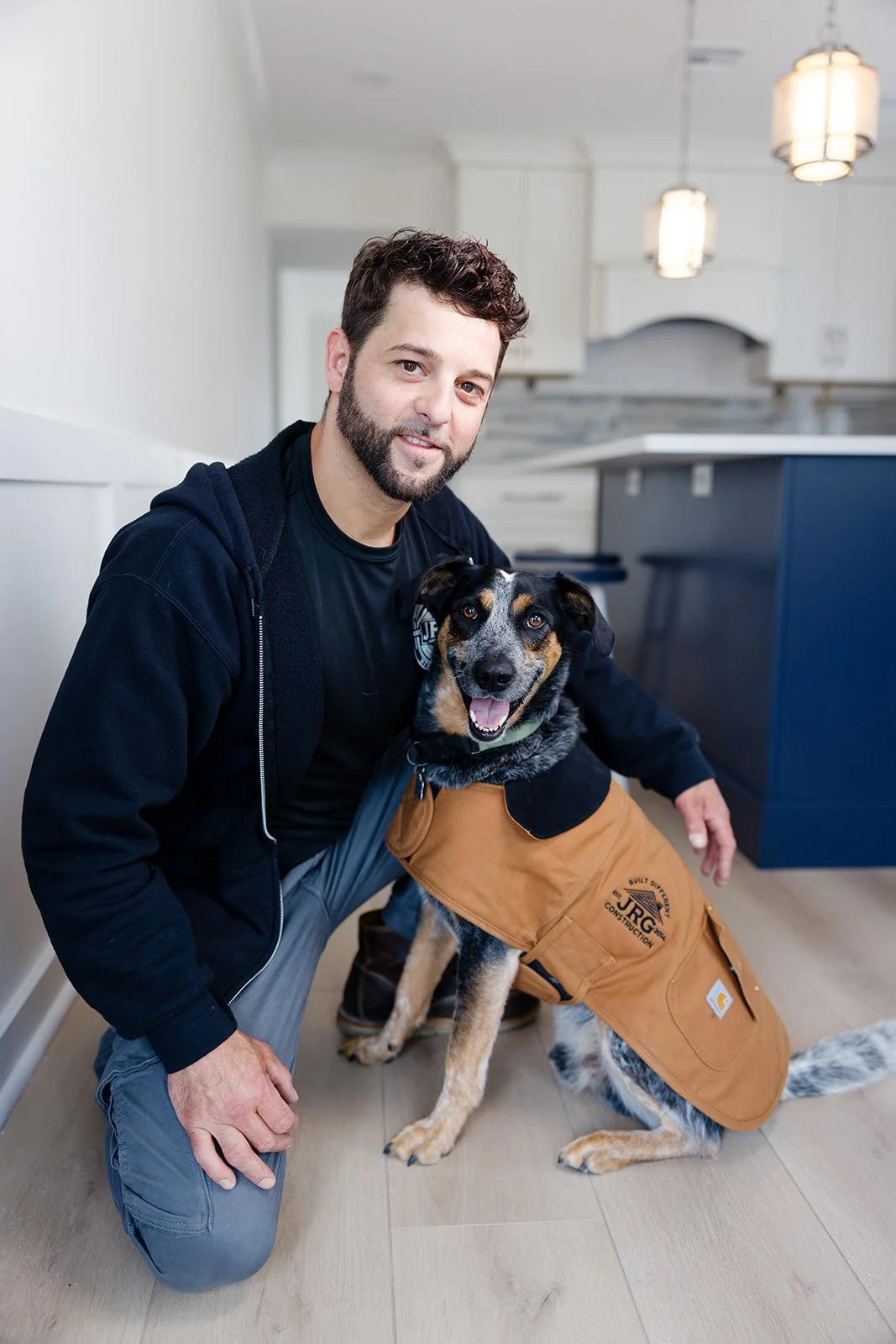 A man kneeling next to a happy service dog with a vest, inside a modern kitchen.