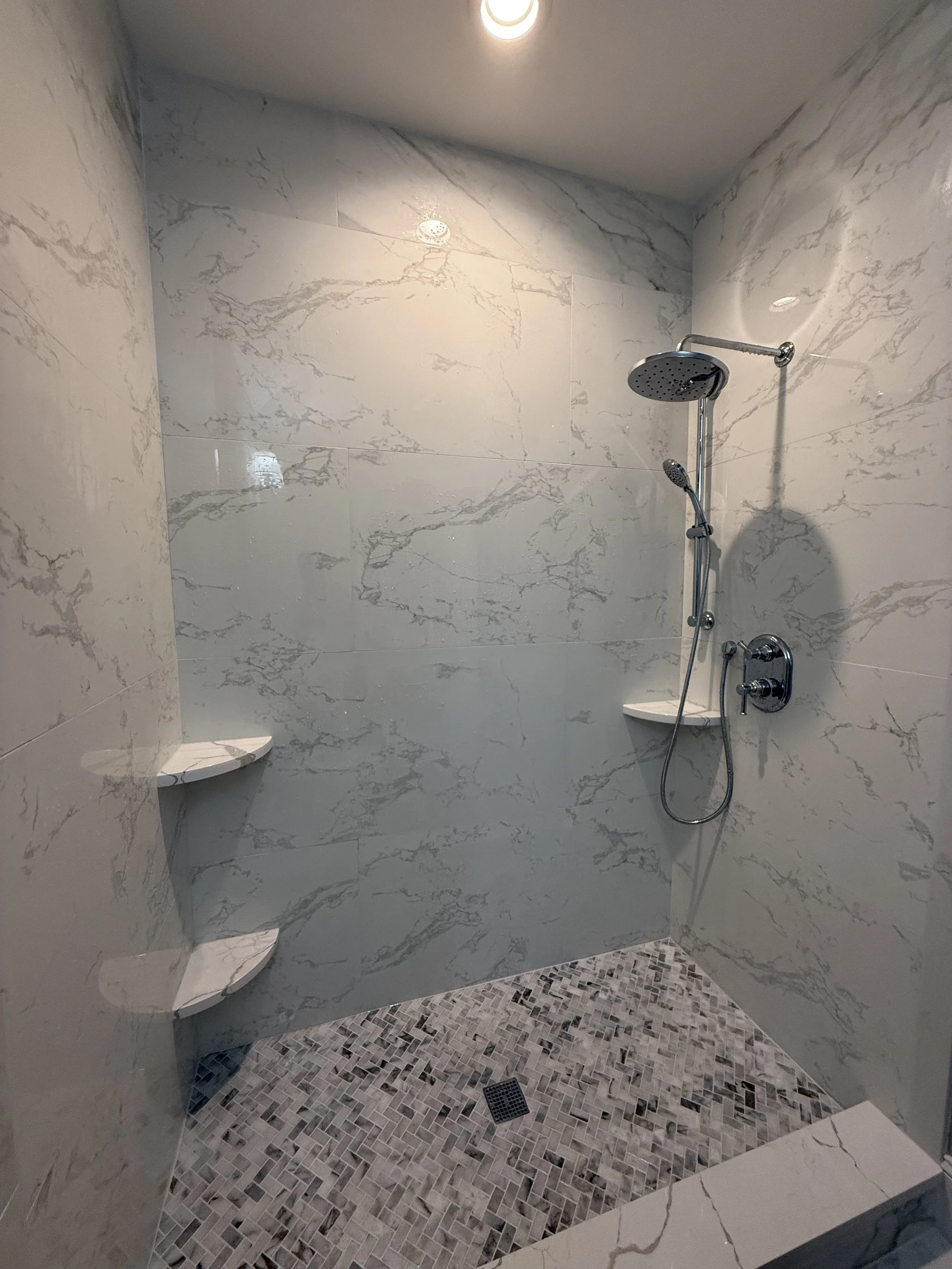 Marble-tiled walk-in shower with a rainfall showerhead, handheld shower, two corner soap niches, and pebble stone floor with drain.