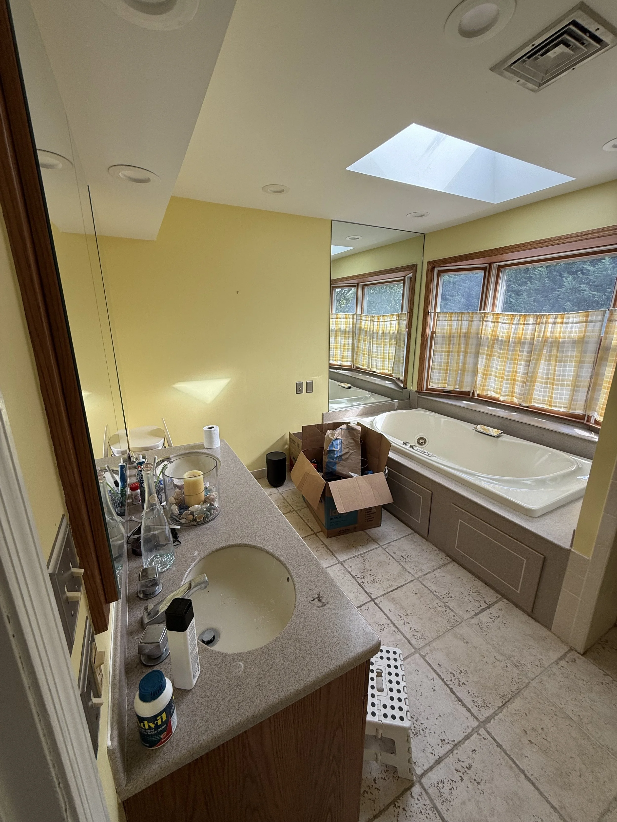 A bathroom with a large bathtub beneath three windows with yellow and white checkered curtains. There is a countertop with various toiletries, a sink, and a mirror. A box filled with items is on the floor near the bathtub, and a step stool is placed in front of the counter.