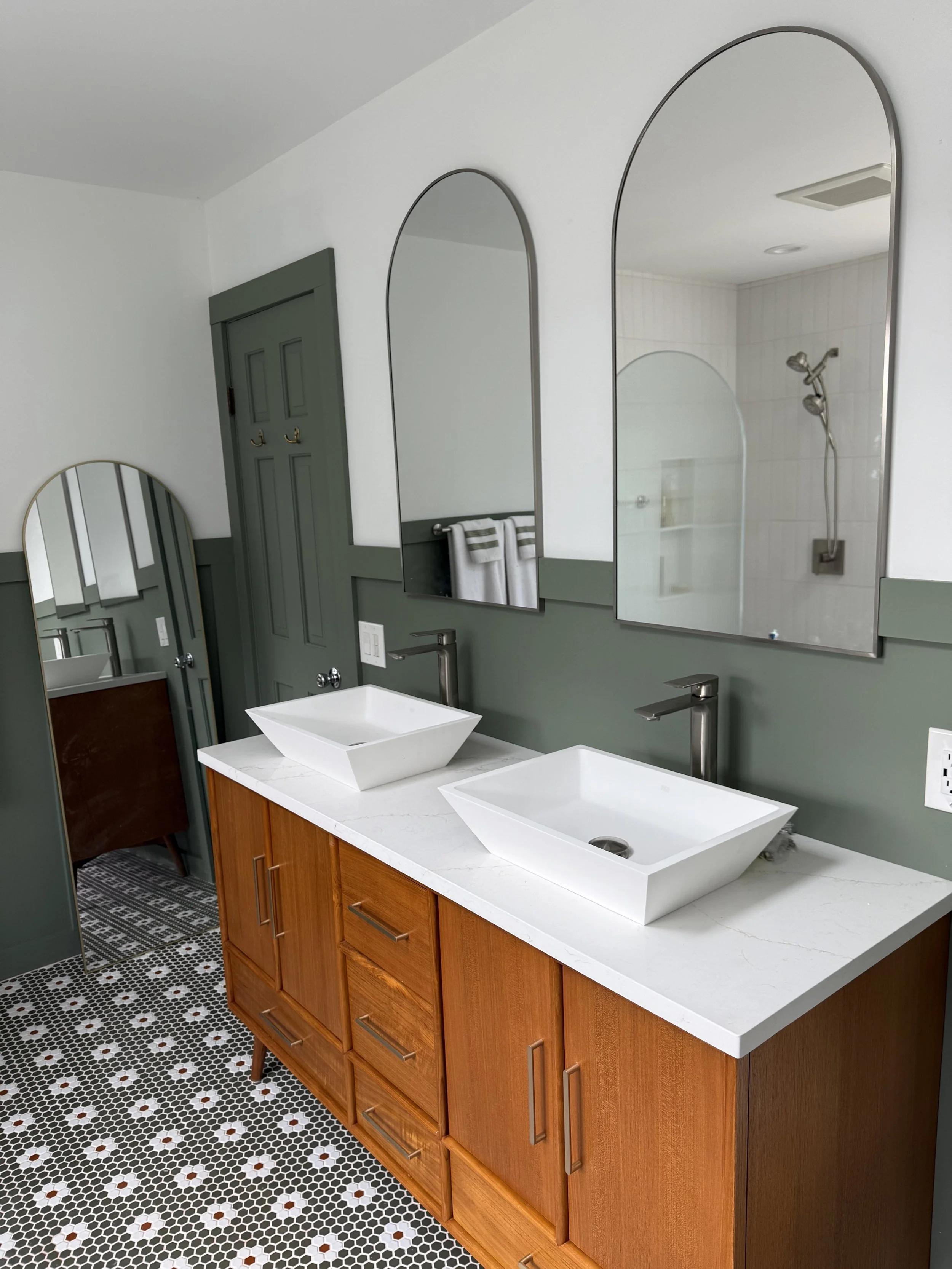 Bathroom vanity with two white vessel sinks, wooden cabinet, large mirrors, a shower with a handheld showerhead, green door, towel, and black and white mosaic floor.