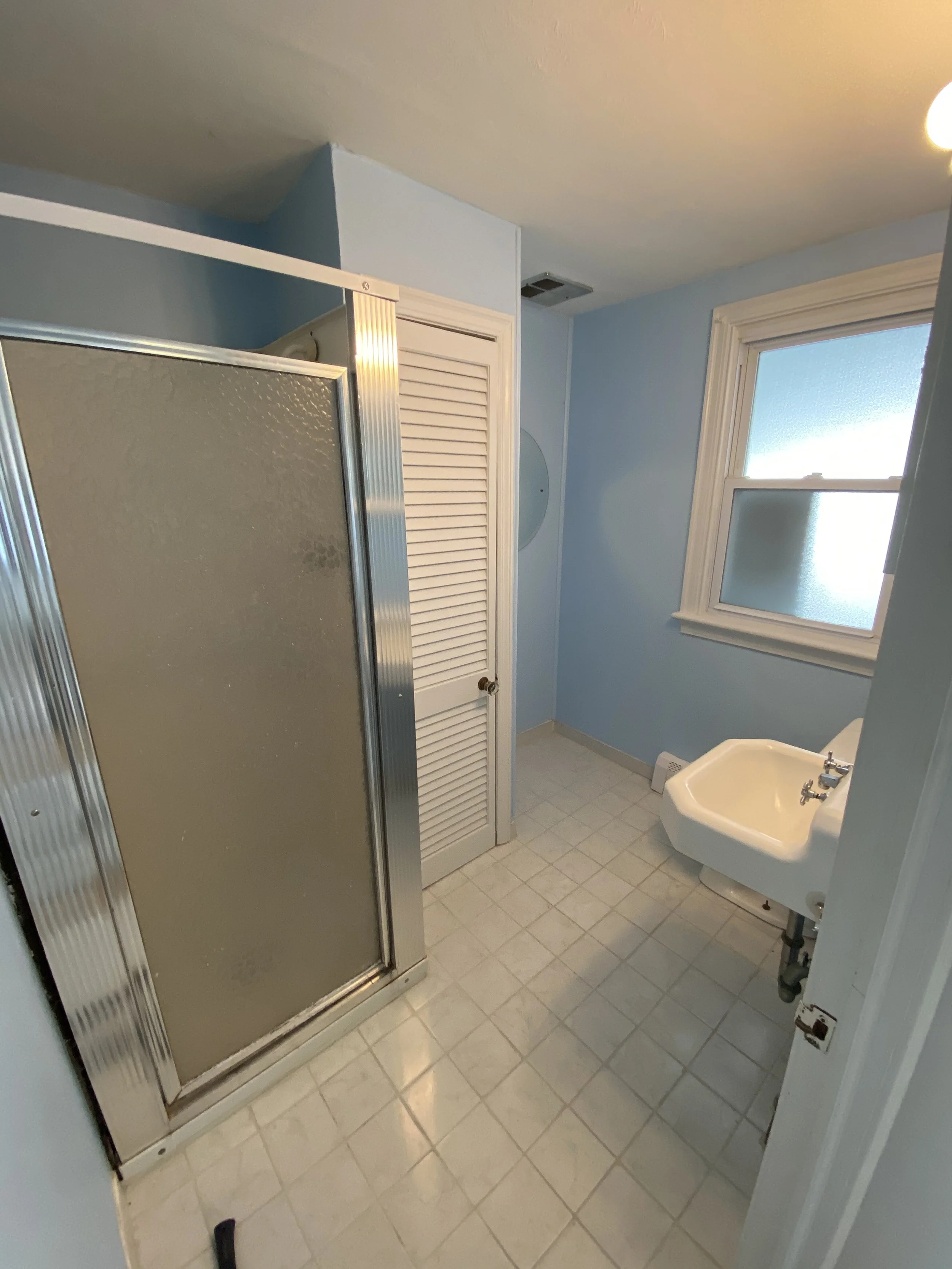 Small bathroom with a frosted window, blue walls, a shower stall with a textured glass door, a white sink, and a louvered closet door.