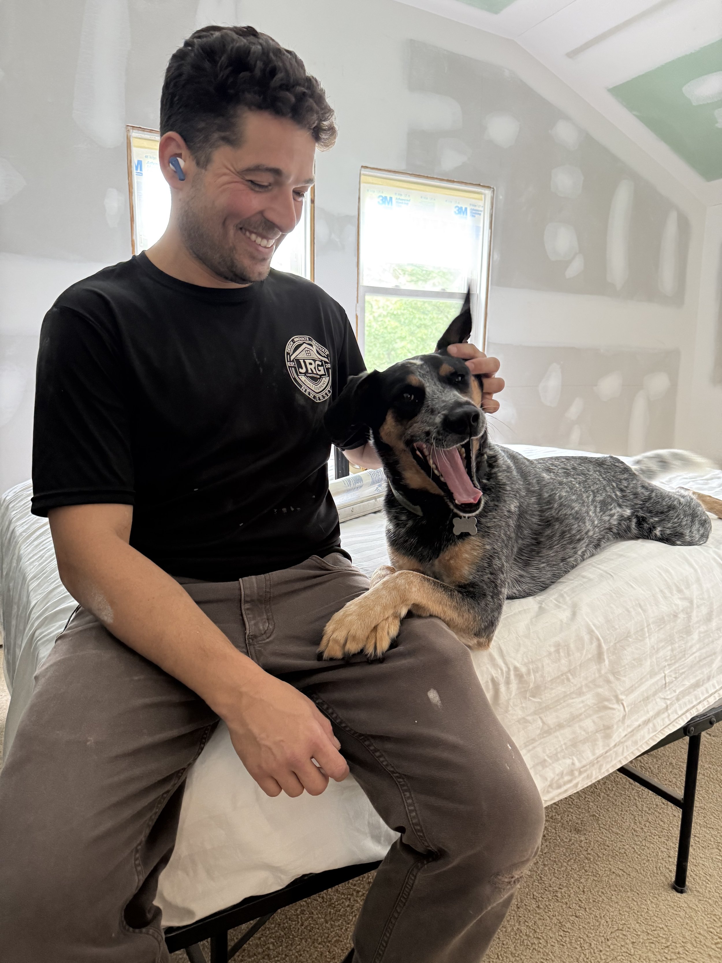 A man with short dark curly hair, wearing a black t-shirt and brown pants, sitting on a bed, smiling and holding a yawning black and tan puppy with a bone-shaped collar. The room has two small windows and unpainted drywall walls.