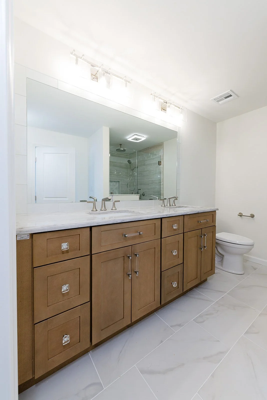 Modern bathroom with wooden vanity, marble countertop, large mirror, and white tiles. Includes a shower with glass door and a toilet.