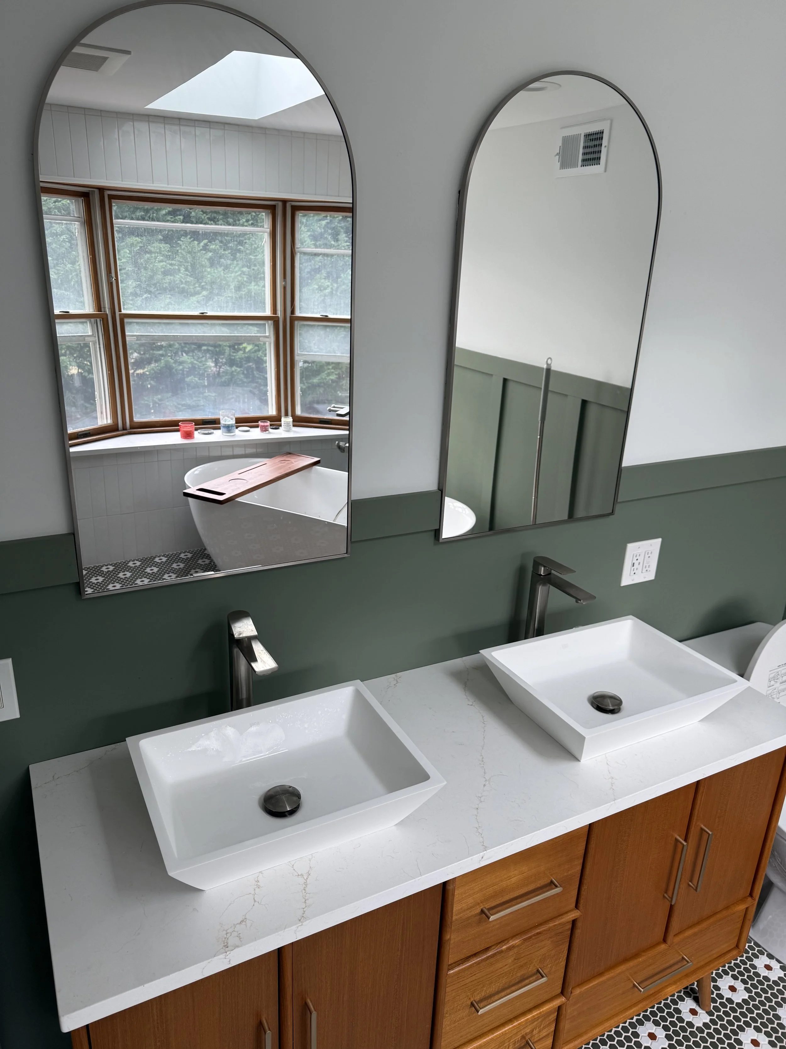A bathroom with two white rectangular vessel sinks on a white marble countertop, wooden vanity cabinets, and two mirrors above the sinks. There is a window with a ledge holding candles and decorative items, and patterned black and white floor tiles.