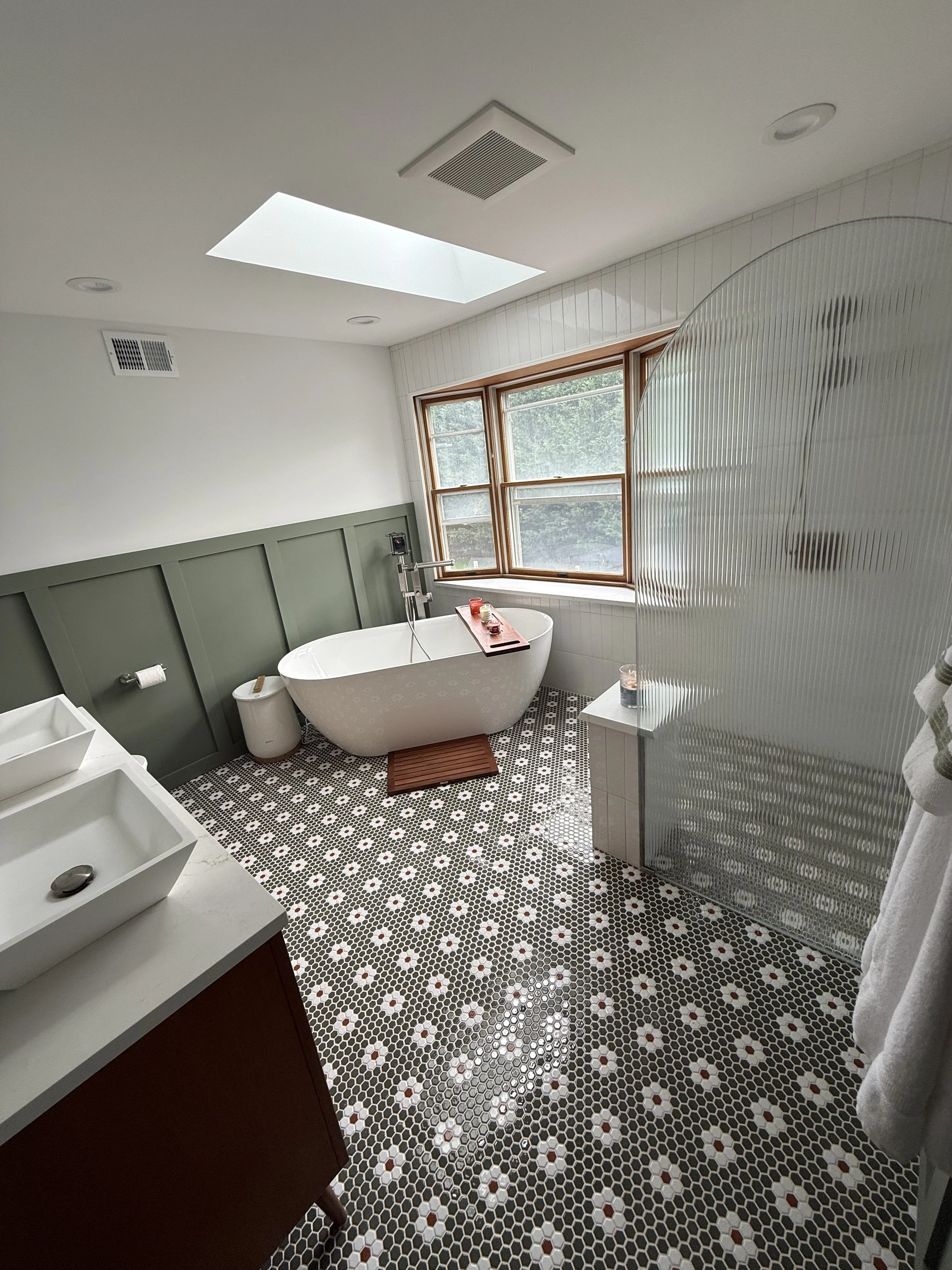 A modern bathroom featuring a freestanding bathtub under a large window, a mosaic tile floor with black, white, and red pattern, a green paneled wall, a white vanity with a vessel sink, and a frosted glass shower screen.