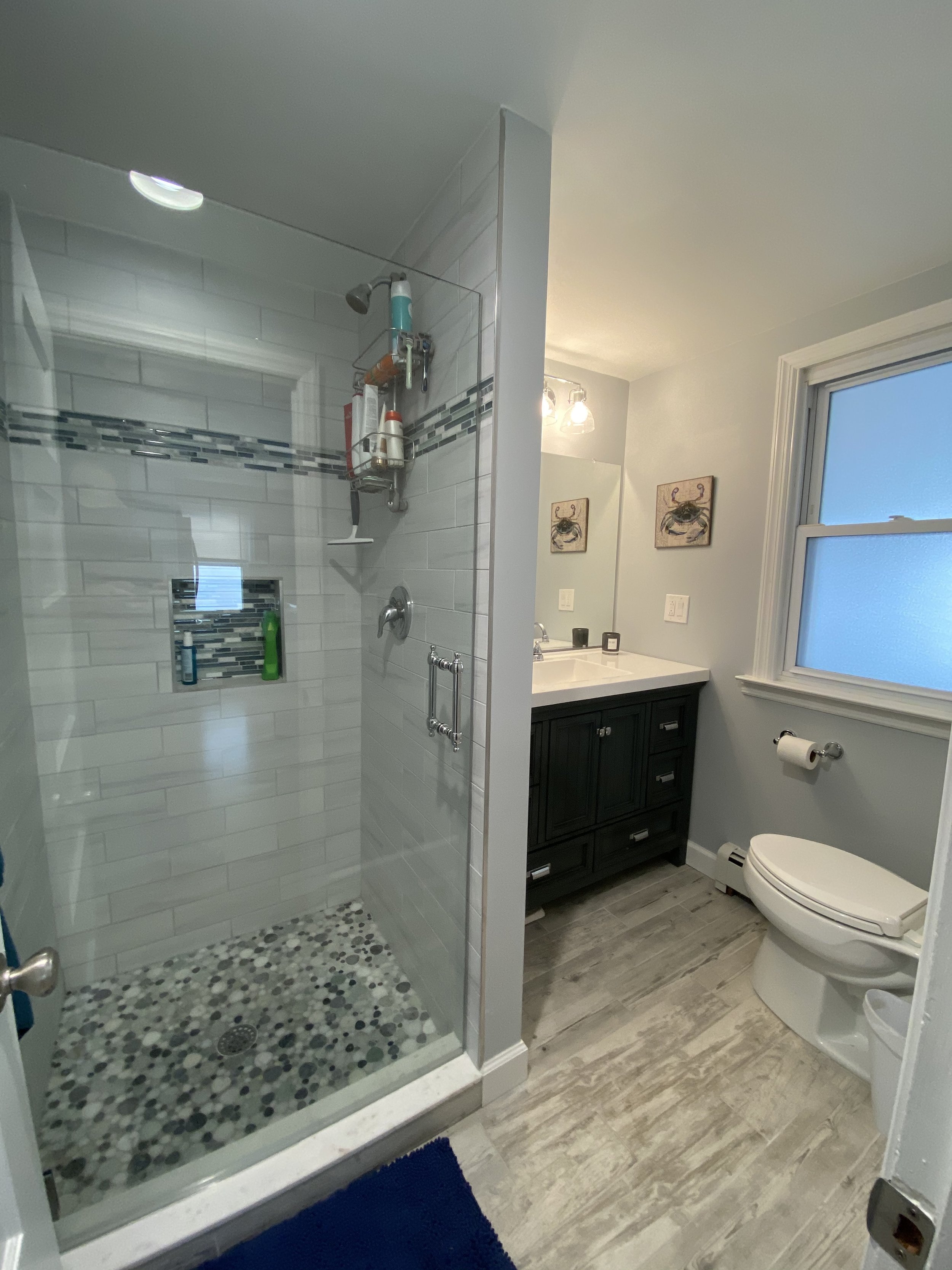Bathroom with a glass shower, white tiles with black and gray accents, a dark vanity with a sink, a mirror, and a window with white trim. There are wall art pictures of crabs, a toilet, a toilet paper holder, and a blue rug.