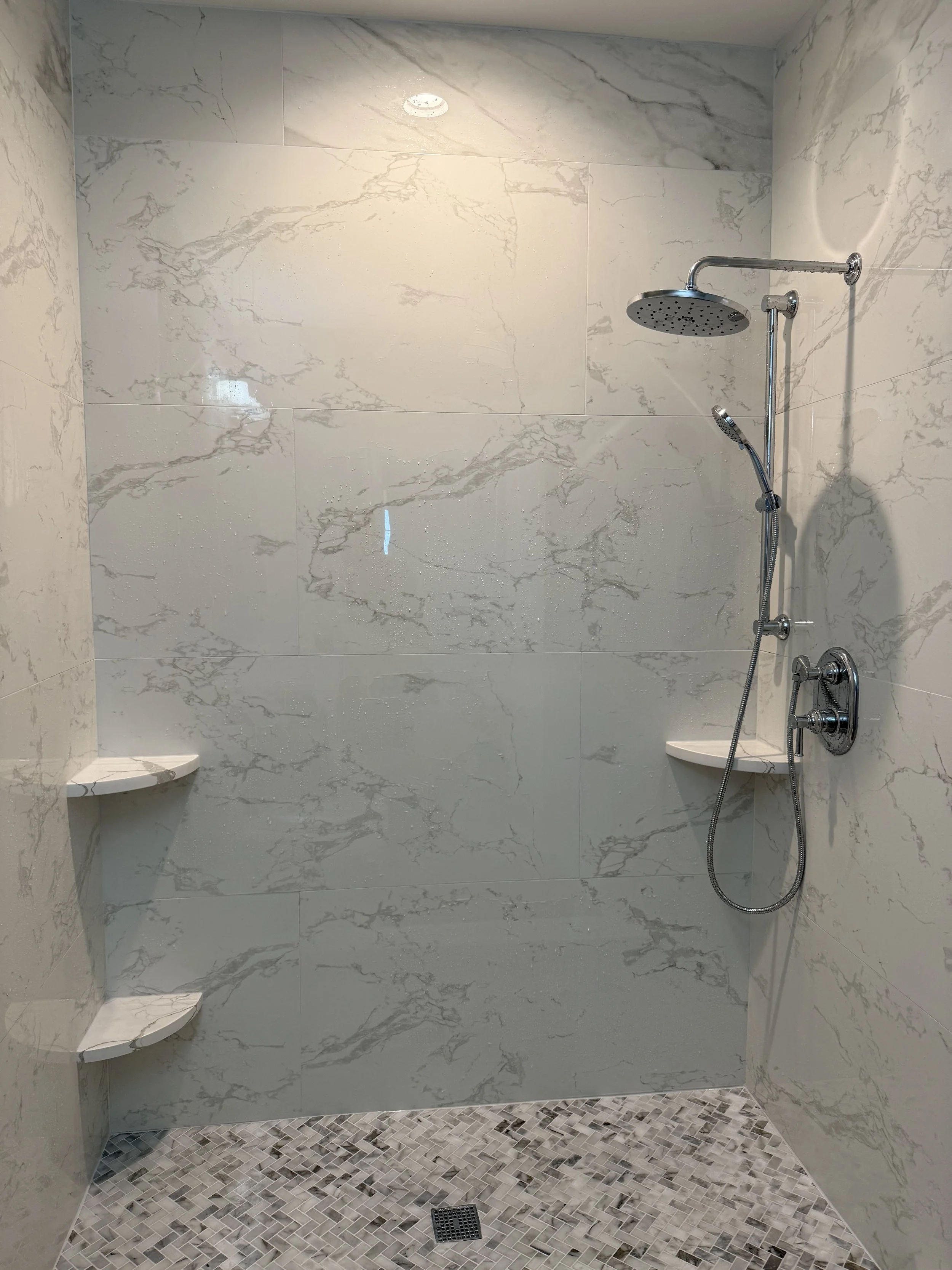 A walk-in shower with white marble tiles, a black circular showerhead, a handheld shower spray, and small mosaic floor tiles. There are small corner shelves on both sides of the shower.