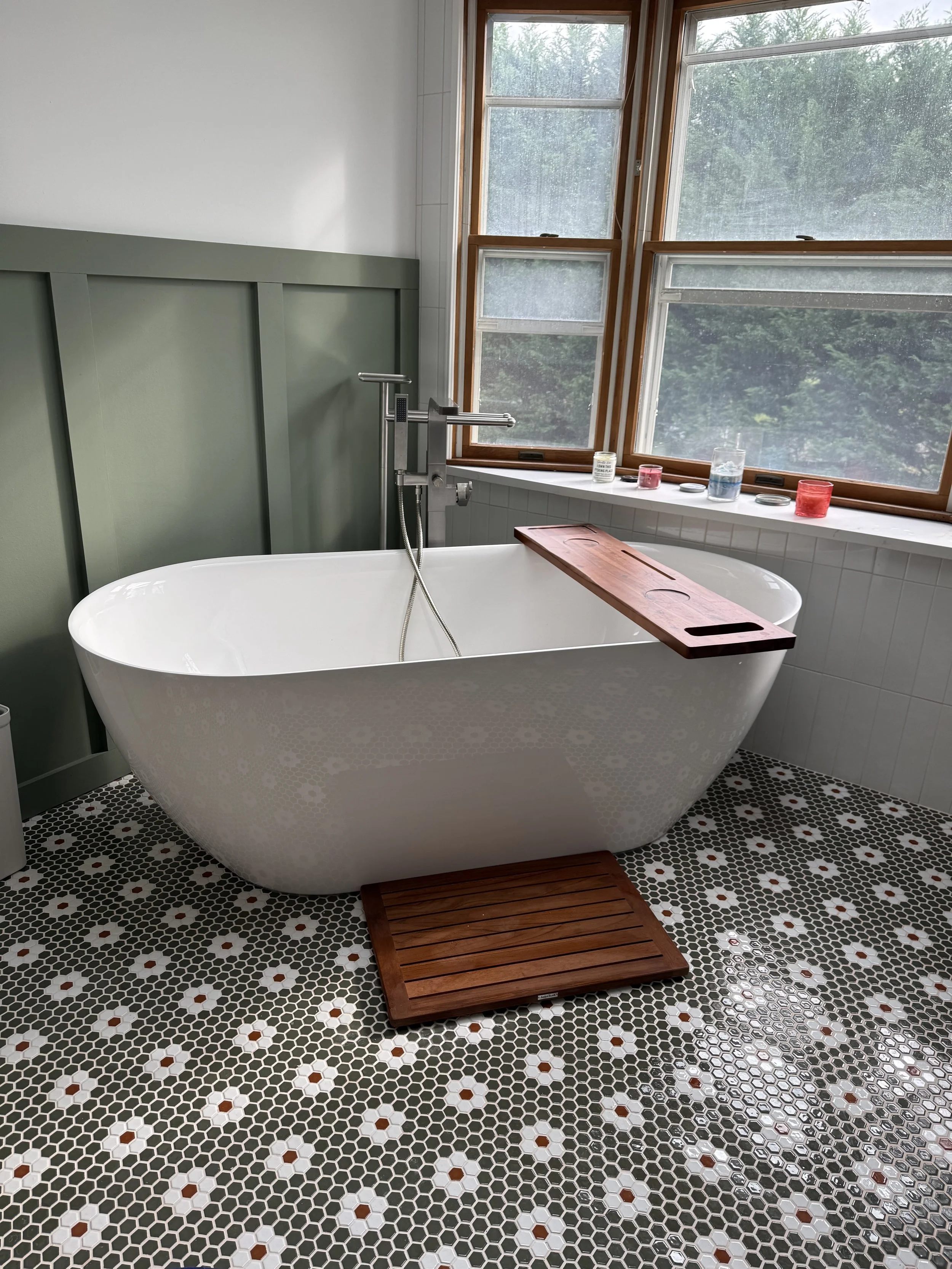 A white freestanding bathtub with a wooden bath tray positioned on a bathroom platform, located below a large window with wooden trim showing greenery outside. The bathroom floor is covered with small hexagonal tiles in black, white, and reddish-brown colors.