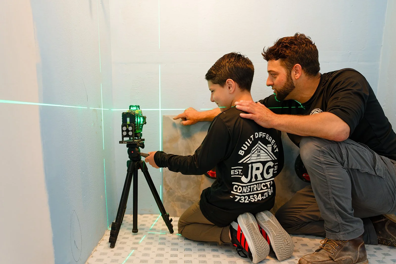 An adult and a boy are kneeling on the floor in front of a wall, using a laser level on a tripod to align with laser lines on the wall. The adult is guiding the boy, who is operating the laser level.
