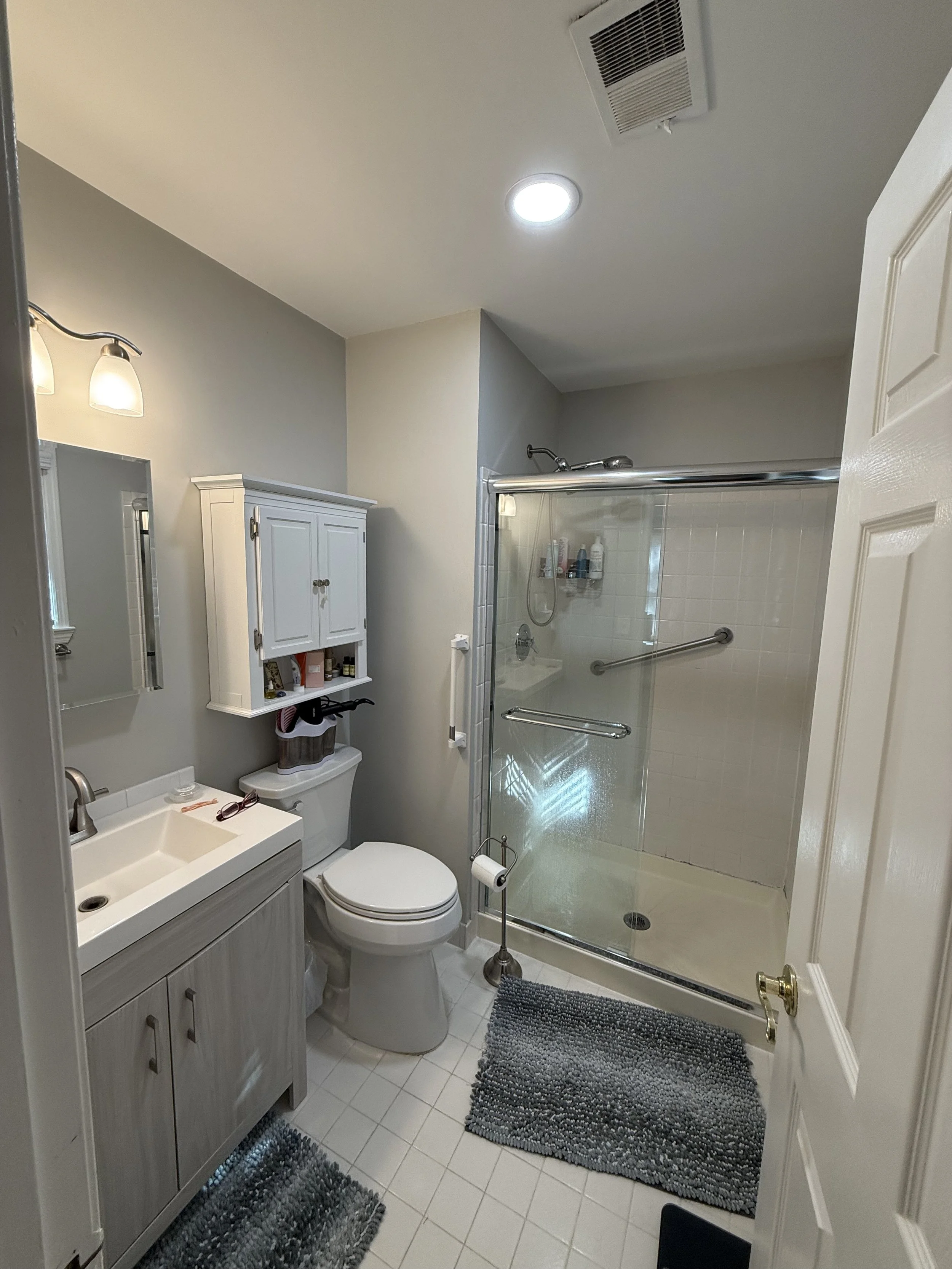 A bathroom with a small sink, mirror, white cabinet, toilet, and glass shower with a sliding door. There are two rugs, one in front of the sink and one in front of the shower, and a toilet paper stand beside the toilet.
