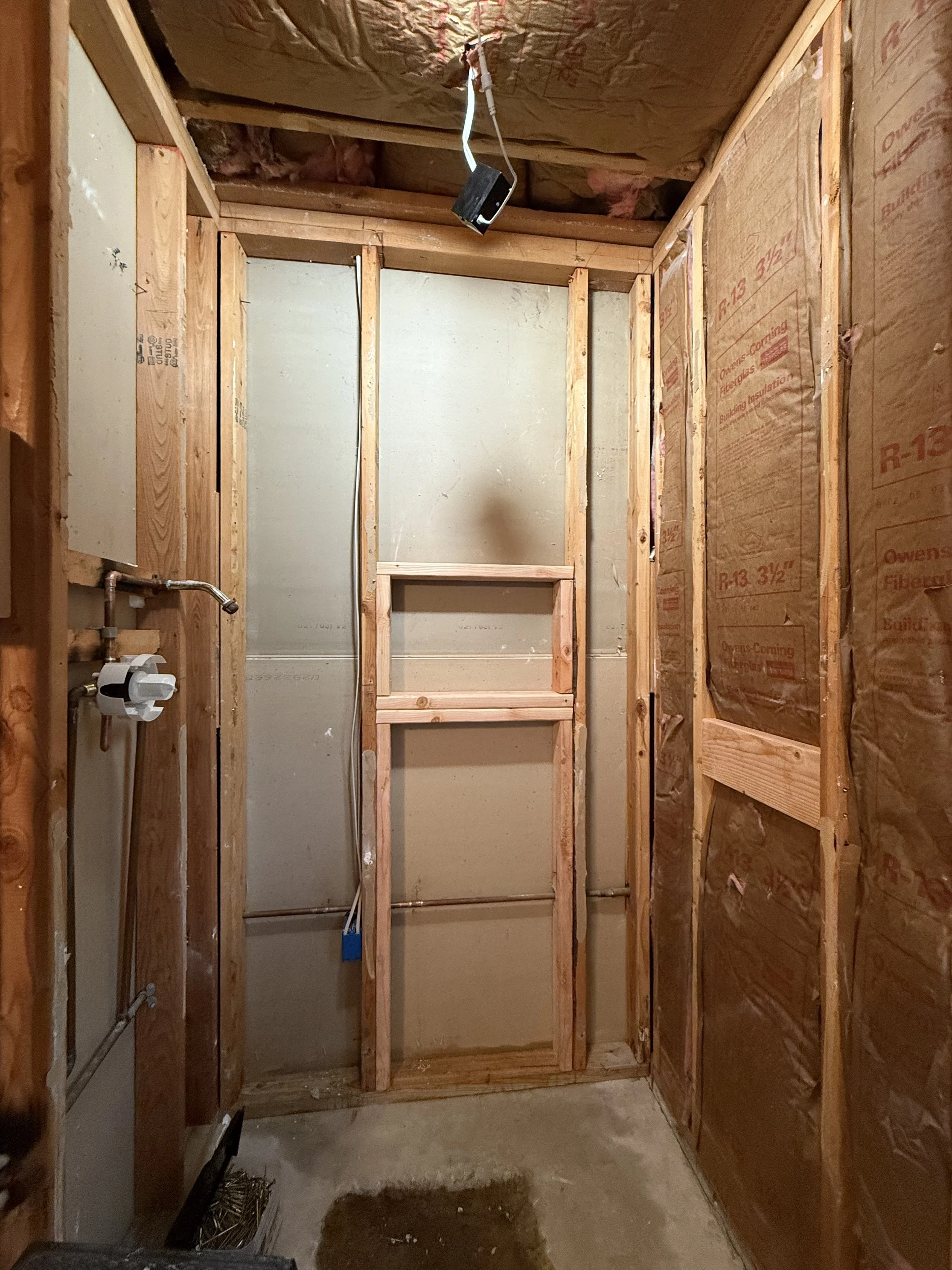 The image shows a small room under construction with exposed wooden framing, insulation on the walls and ceiling, and rough plumbing and wiring still visible.