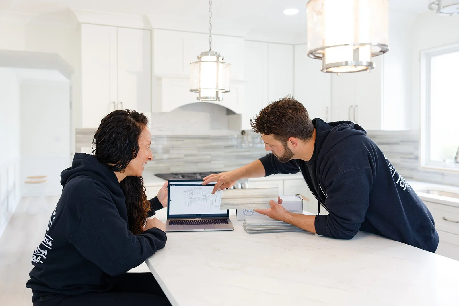 Two people in black hoodies discuss kitchen design plans at a white kitchen island, with a laptop displaying a kitchen layout and several samples of material or paint on the table.