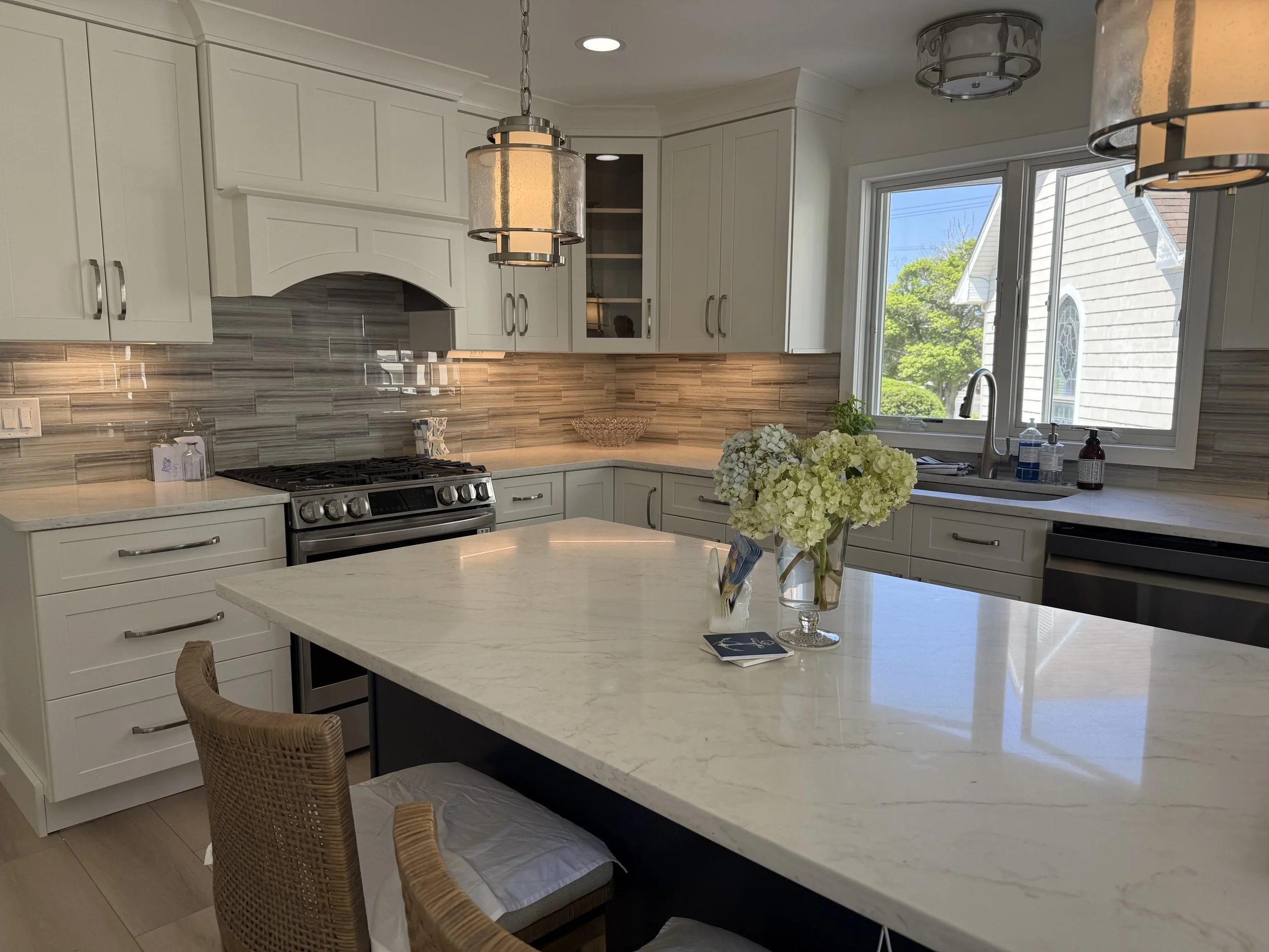 Modern kitchen with white cabinets, a marble island, and pendant lights, with a window showing a sunny exterior with green trees.