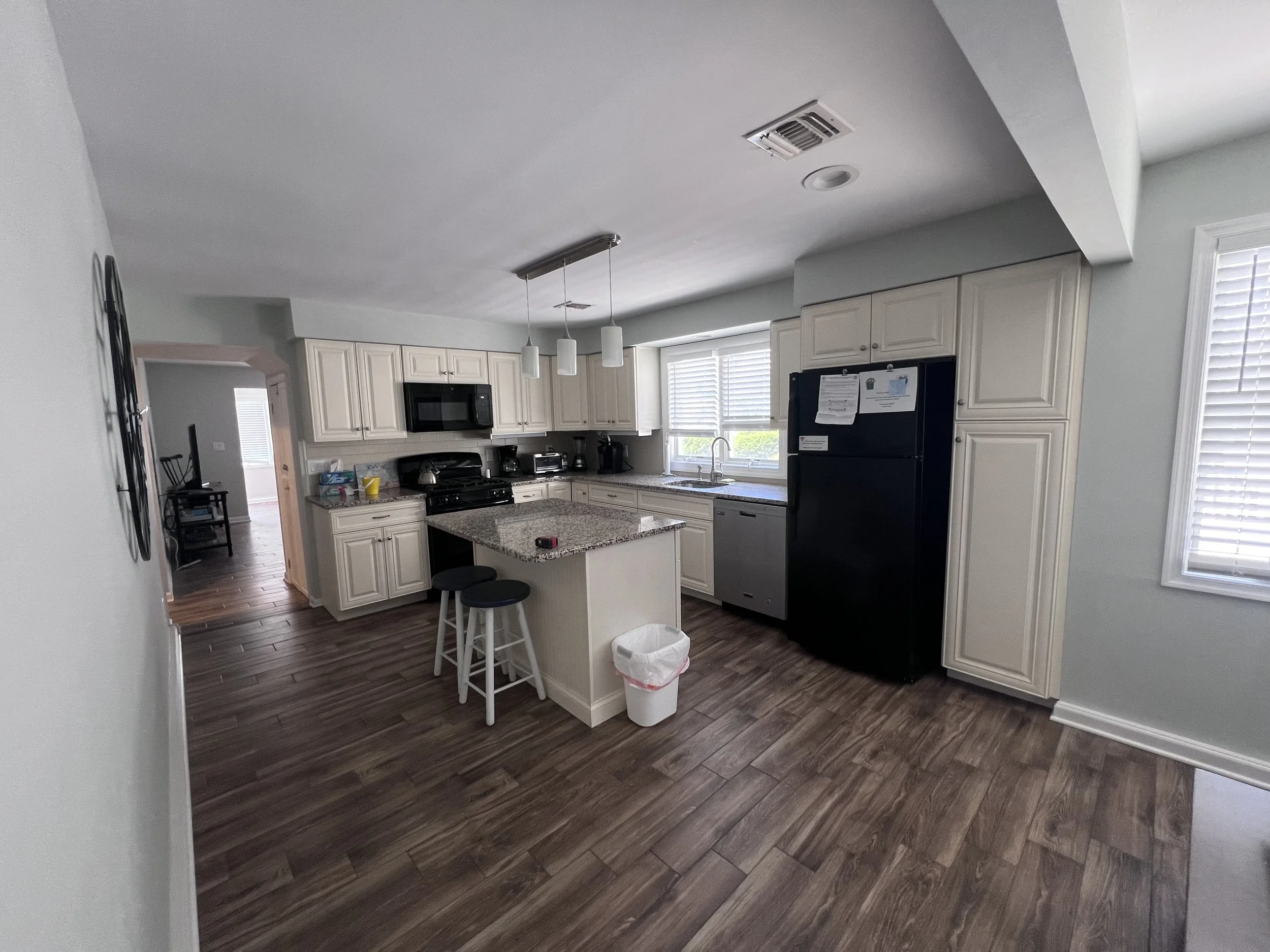 View of a kitchen with white cabinets, granite countertops, a black refrigerator, a microwave, a stove, and a small island with two stools. There are windows with blinds and wood flooring.